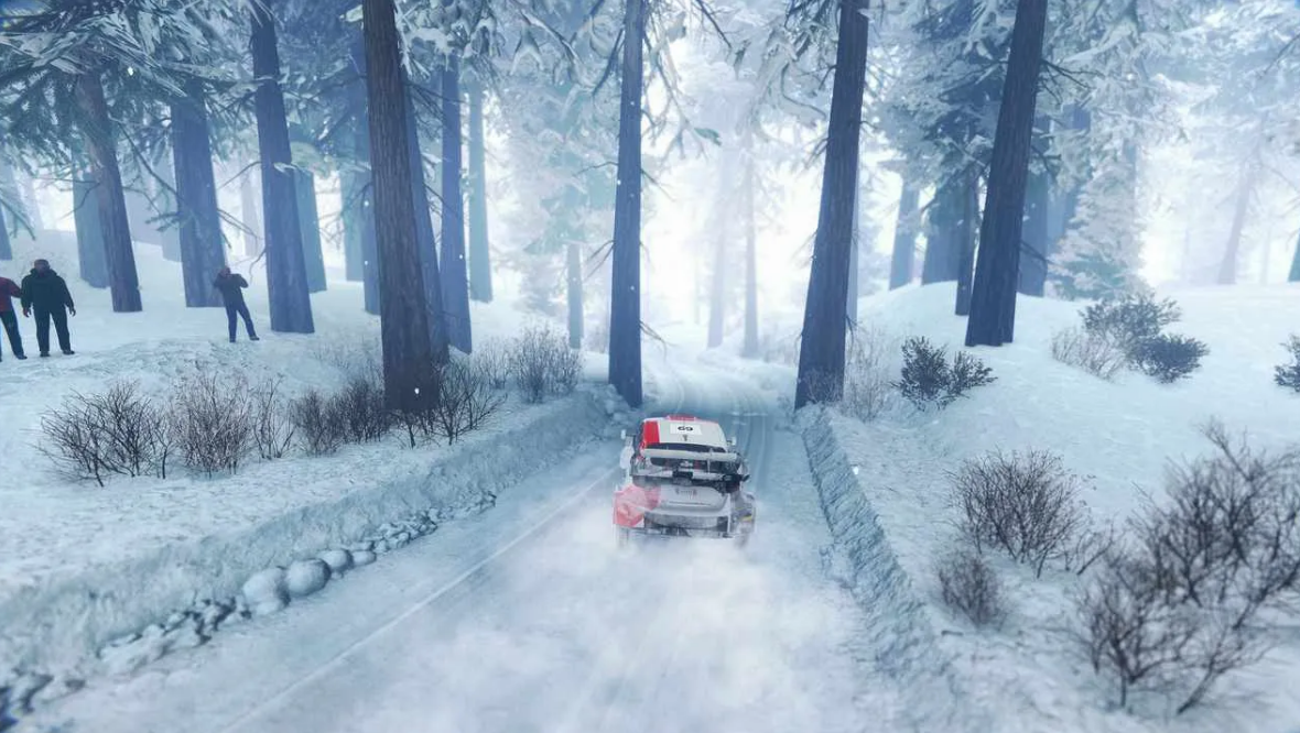 Ein Auto fährt auf einer schneebedeckten Straße in einem Wald. Bäume säumen die Straße, links stehen Menschen.