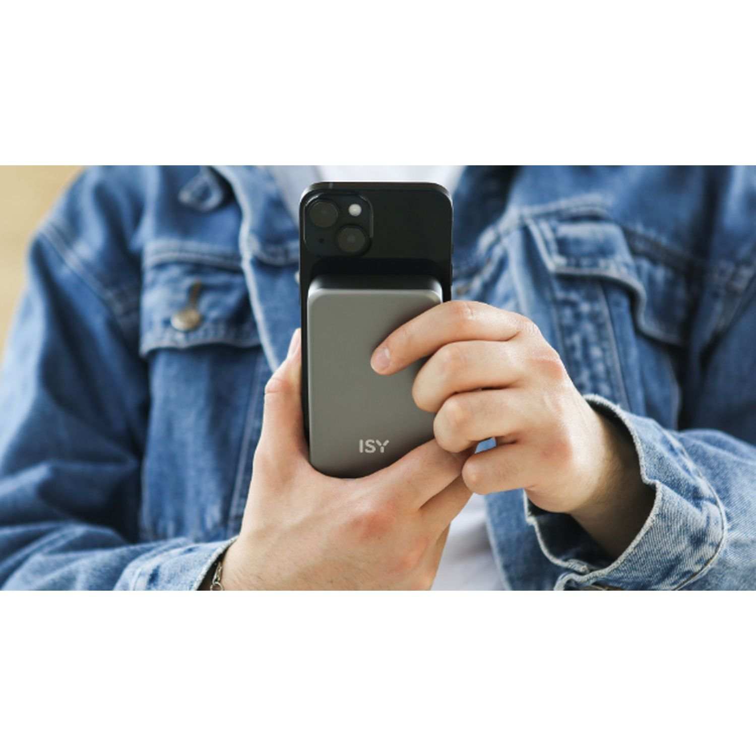 Hands holding a phone with a silver device attached to it. The person wears a denim jacket.