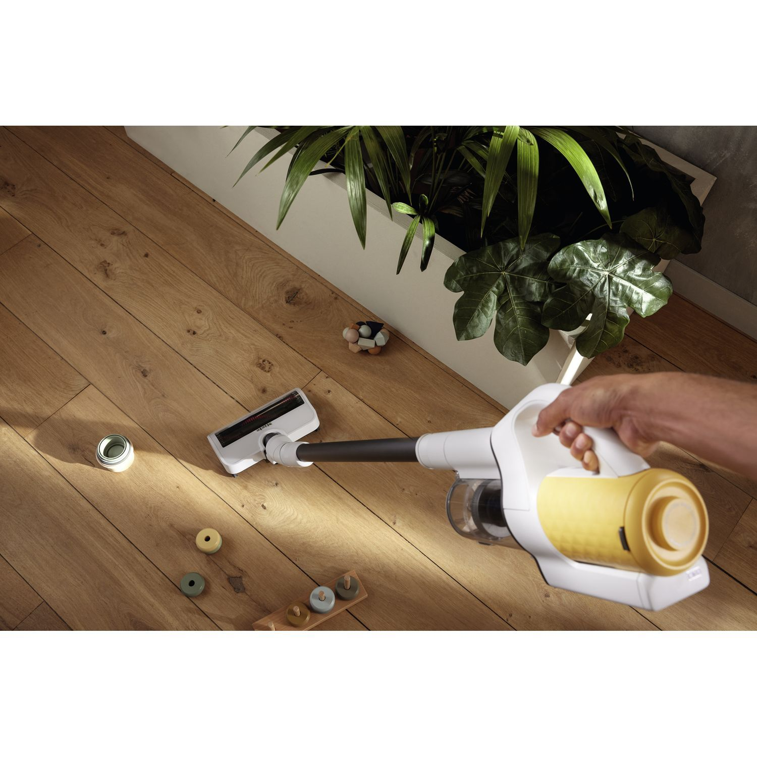 Person using a white and yellow vacuum cleaner on wooden flooring. Plants and toys are visible.