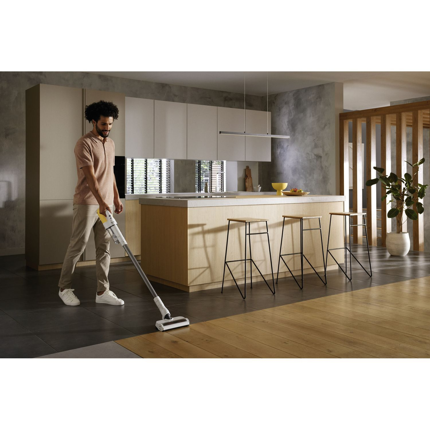 Man vacuuming a wooden floor. A kitchen island, stools, and a plant are in the background.