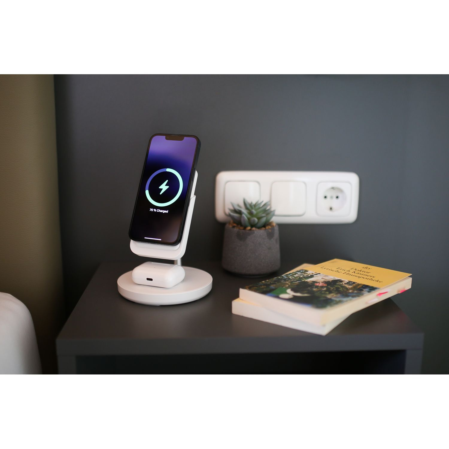 Téléphone en charge sur un chargeur sans fil blanc. Une plante et un livre sont à proximité. Fond gris.