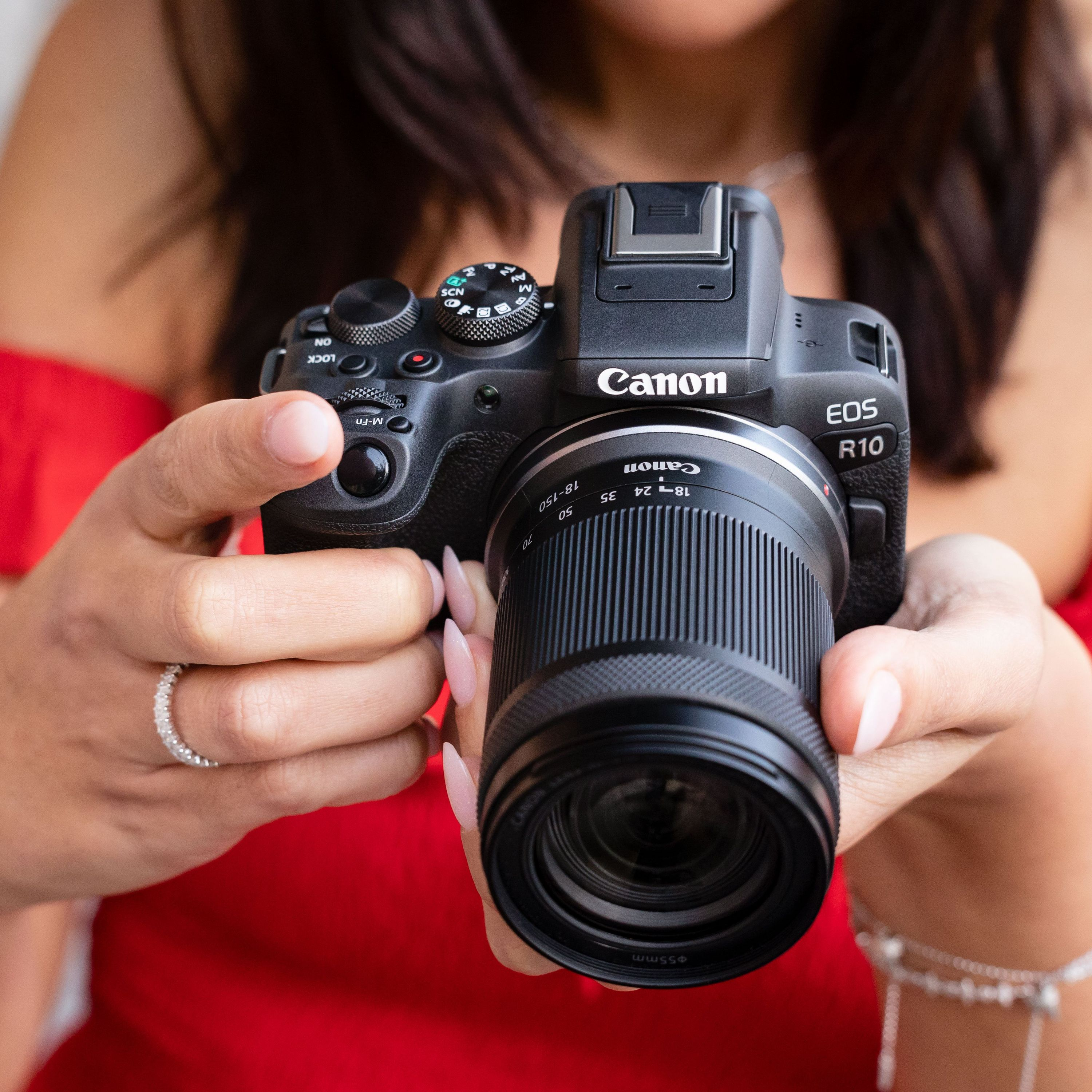 Une femme tient un appareil photo Canon EOS R10, avec un objectif, devant un fond rouge.