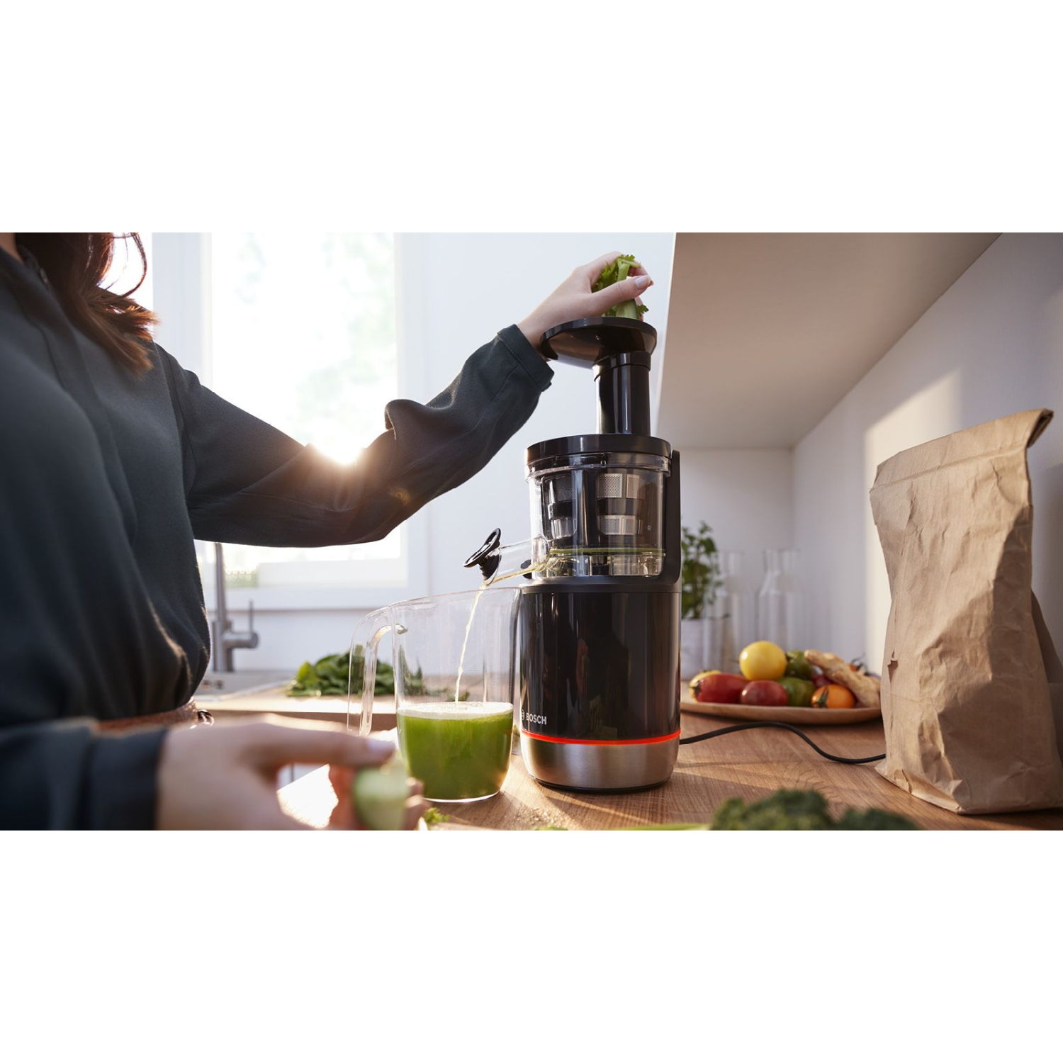 Een vrouw die groenten toevoegt aan een juicer met een glas sap op het aanrecht.