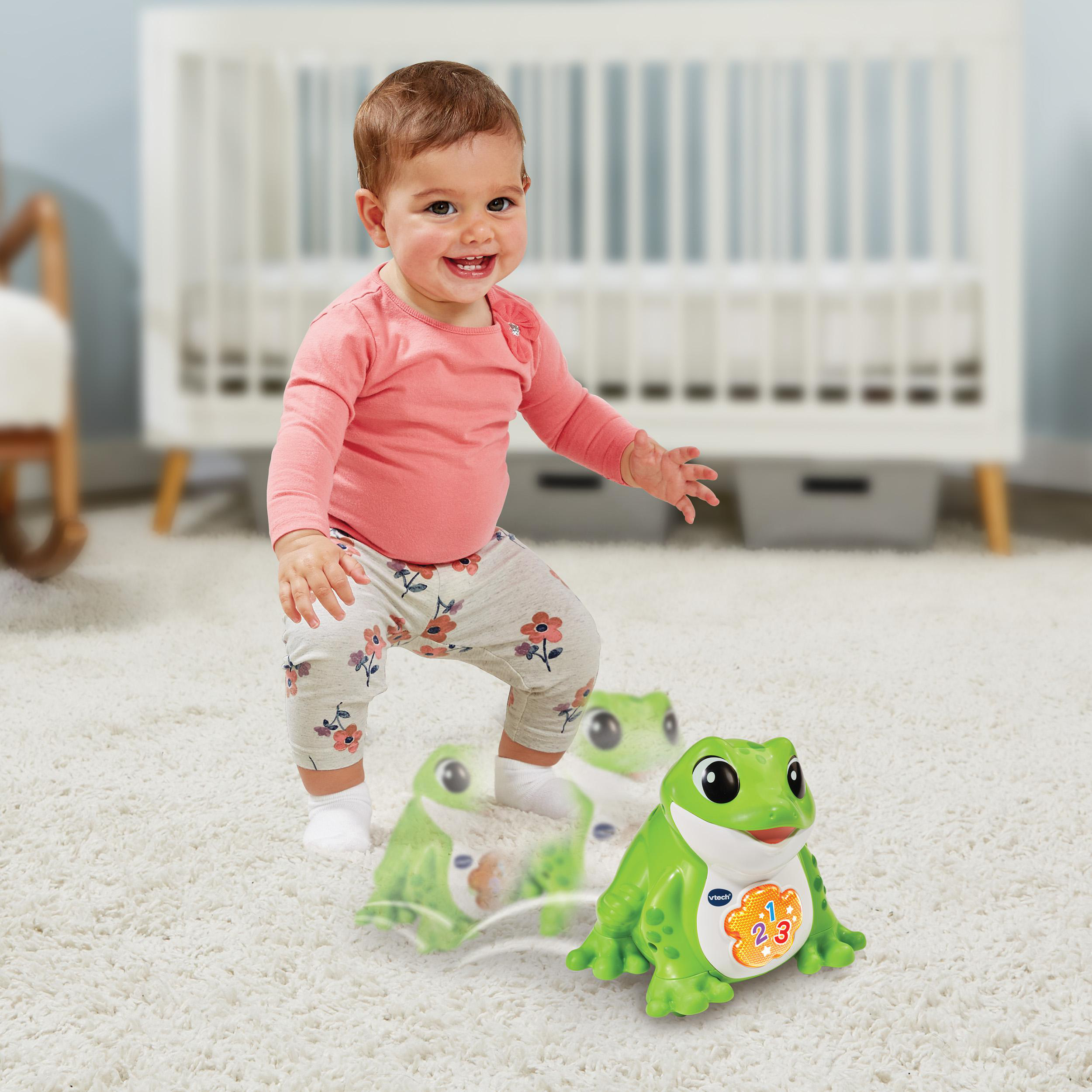 Ein Baby lächelt und steht in der Nähe grüner Spielzeugfrösche auf einem weißen Teppich, in einem hellen Raum mit einer Krippe im Hintergrund.