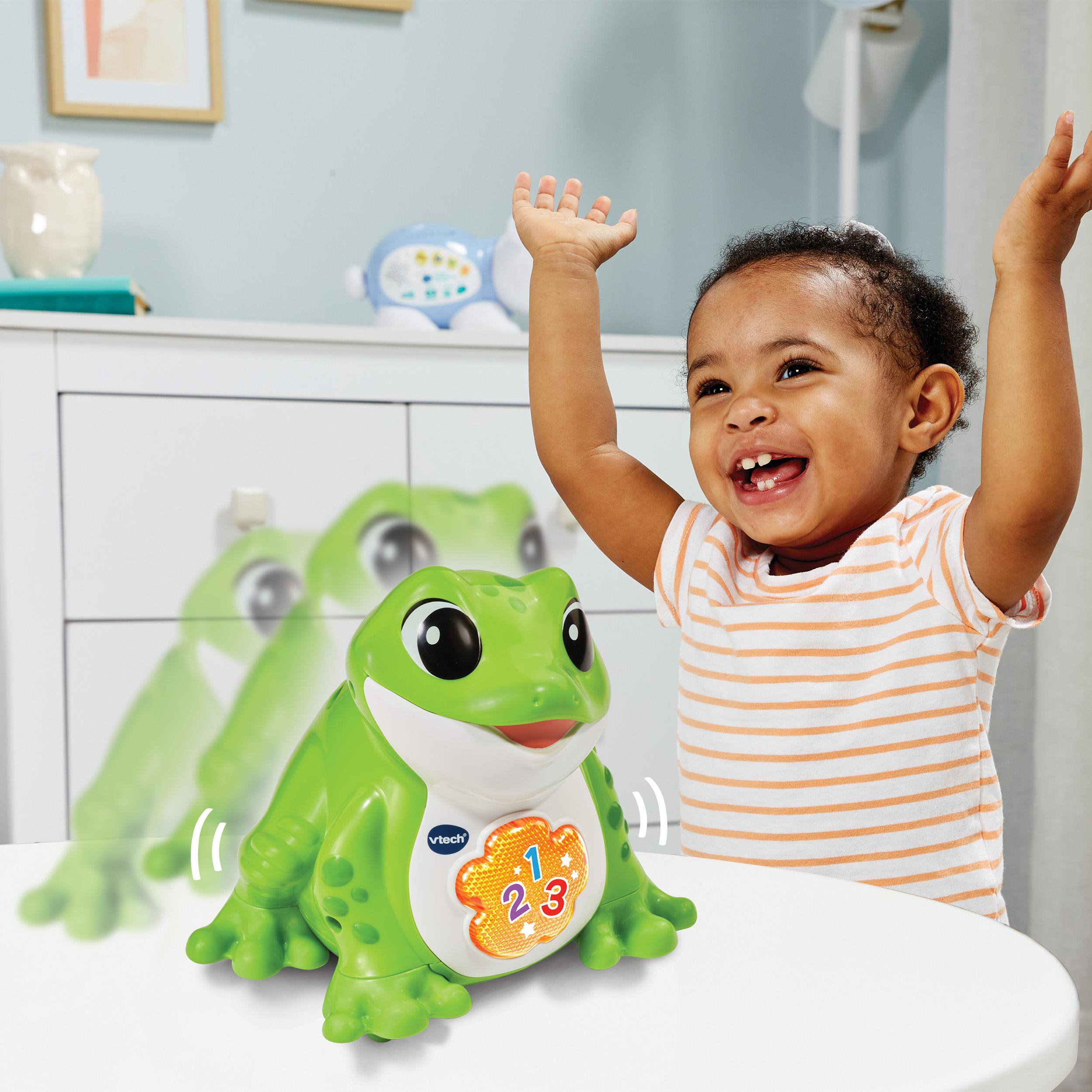 Ein Kind lächelt einen grünen Frosch an. Das Spielzeug befindet sich auf einer weißen Oberfläche.