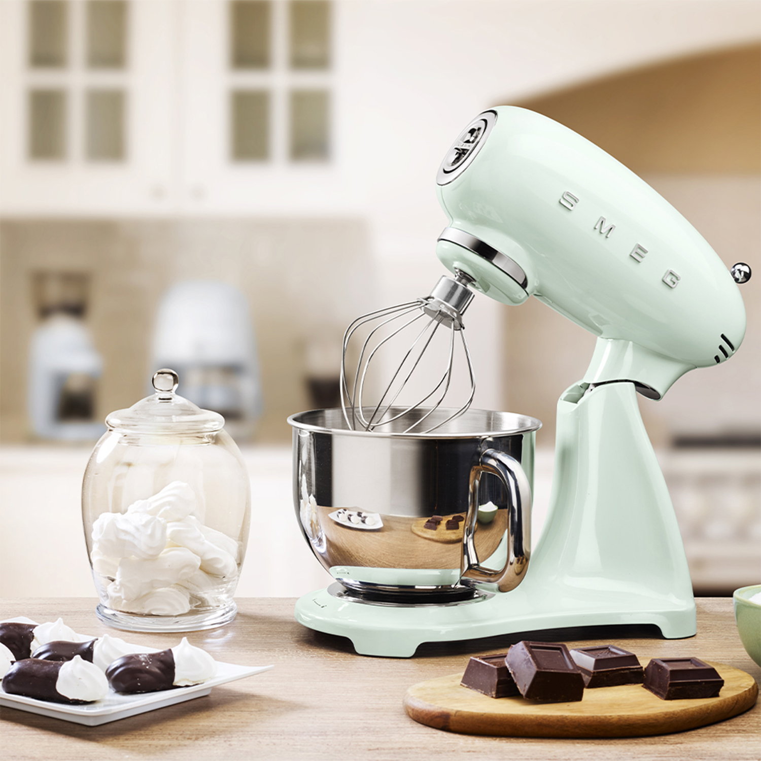 Un mixer Smeg verde menta con accessori su un tavolo di legno, con cibi e sfondo sfocato.
