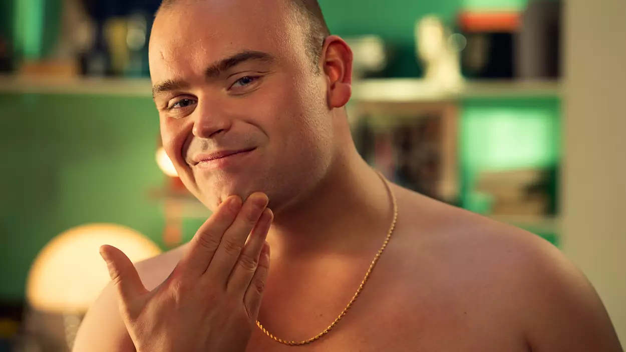 Un homme avec un collier en or se touche le visage, souriant. Il est torse nu et le fond est flou.
