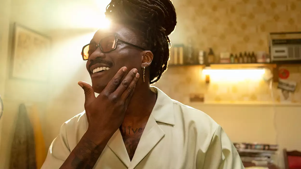 Un homme avec des dreadlocks, portant des lunettes, souriant, touchant son visage, dans une pièce.