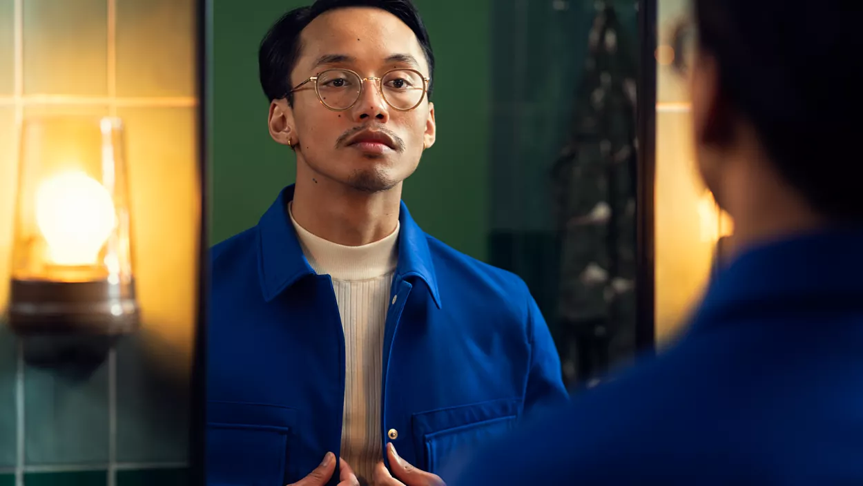 Un homme en veste bleue regarde son reflet dans le miroir d'une salle de bain.