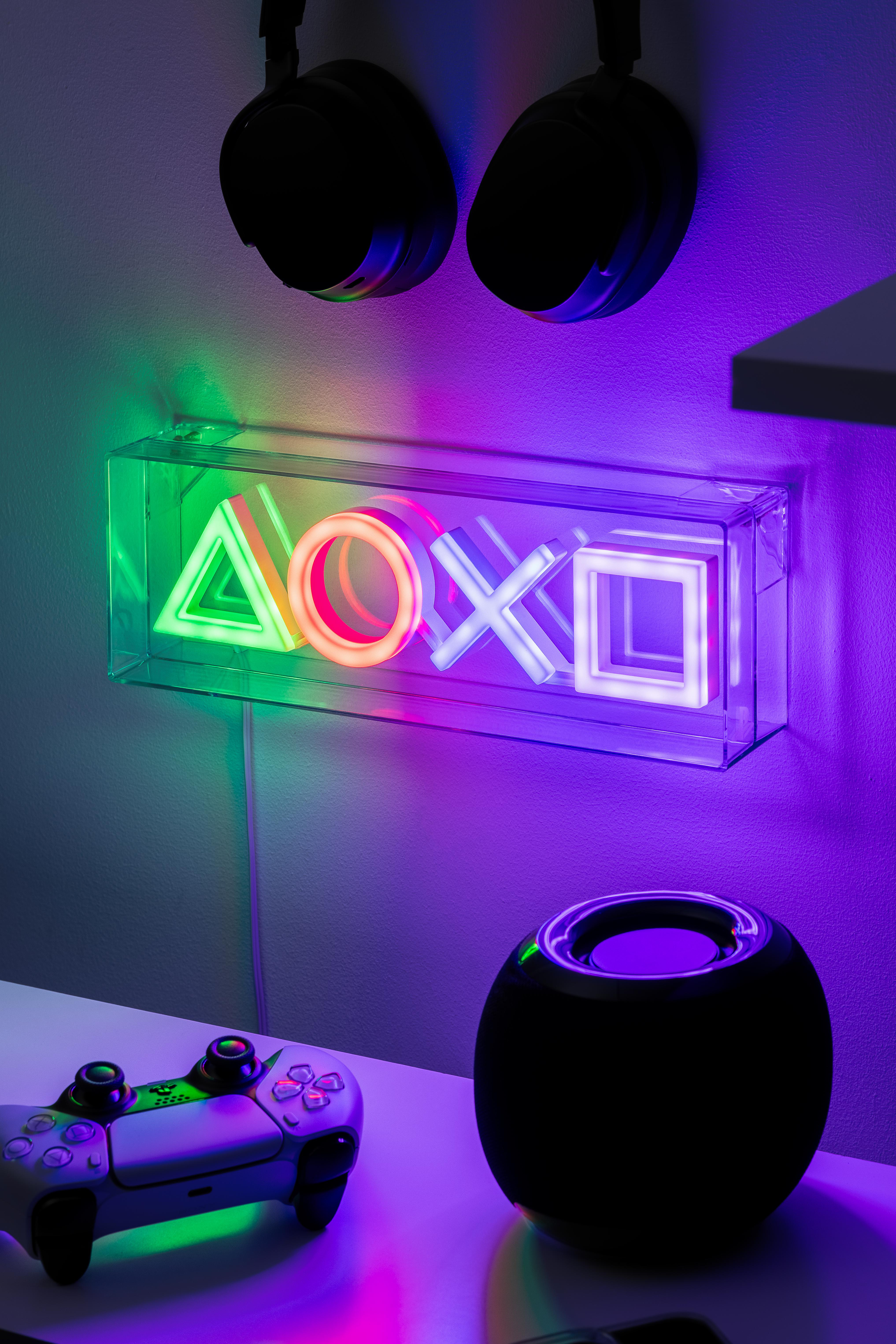 PlayStation-Controller und Neon-Schild mit PlayStation-Symbolen an einer Wand. Kopfhörer hängen oben.