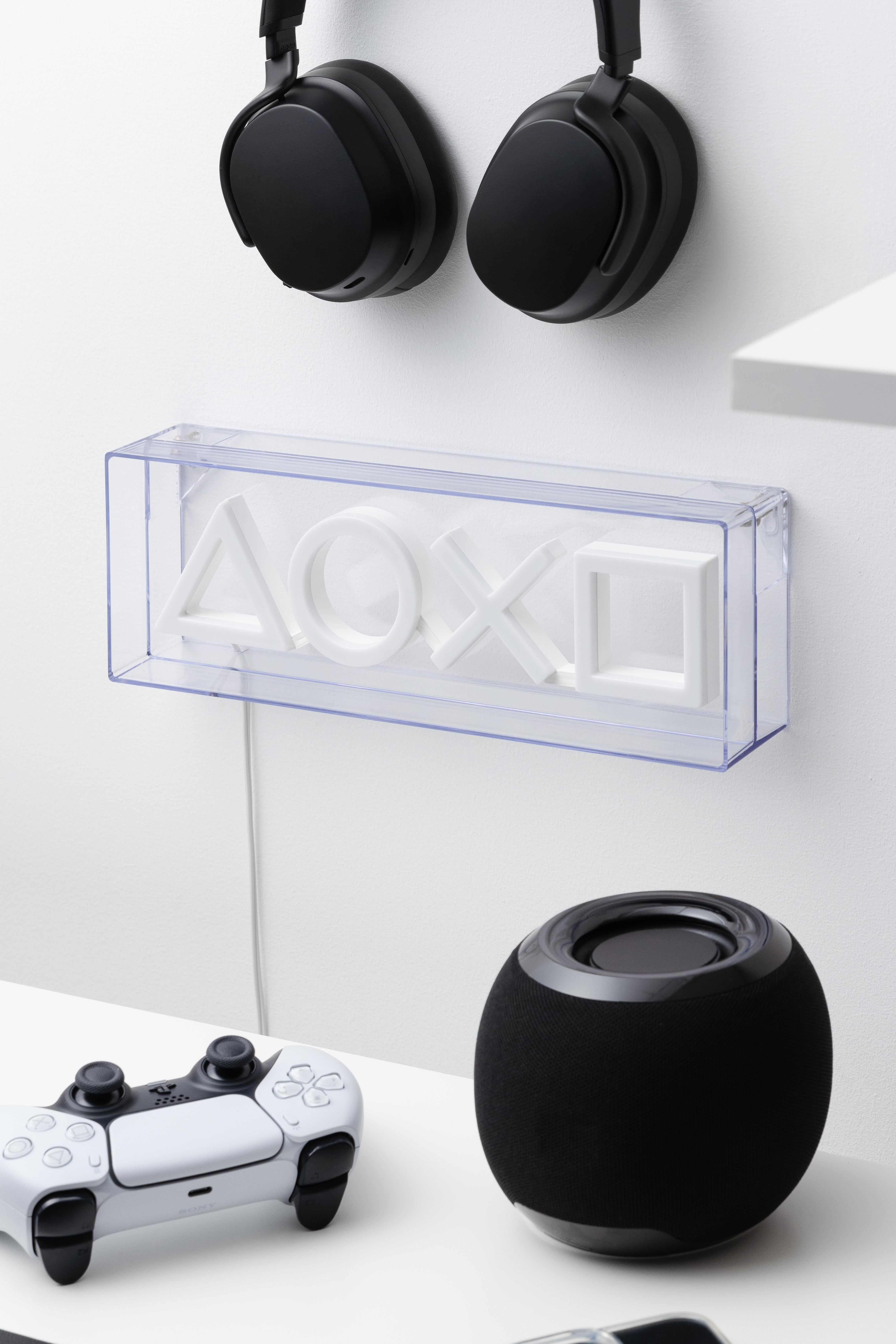 Weiße Wand mit transparenter Box mit Playstation-Icons und Kopfhörern. Auch ein Lautsprecher.