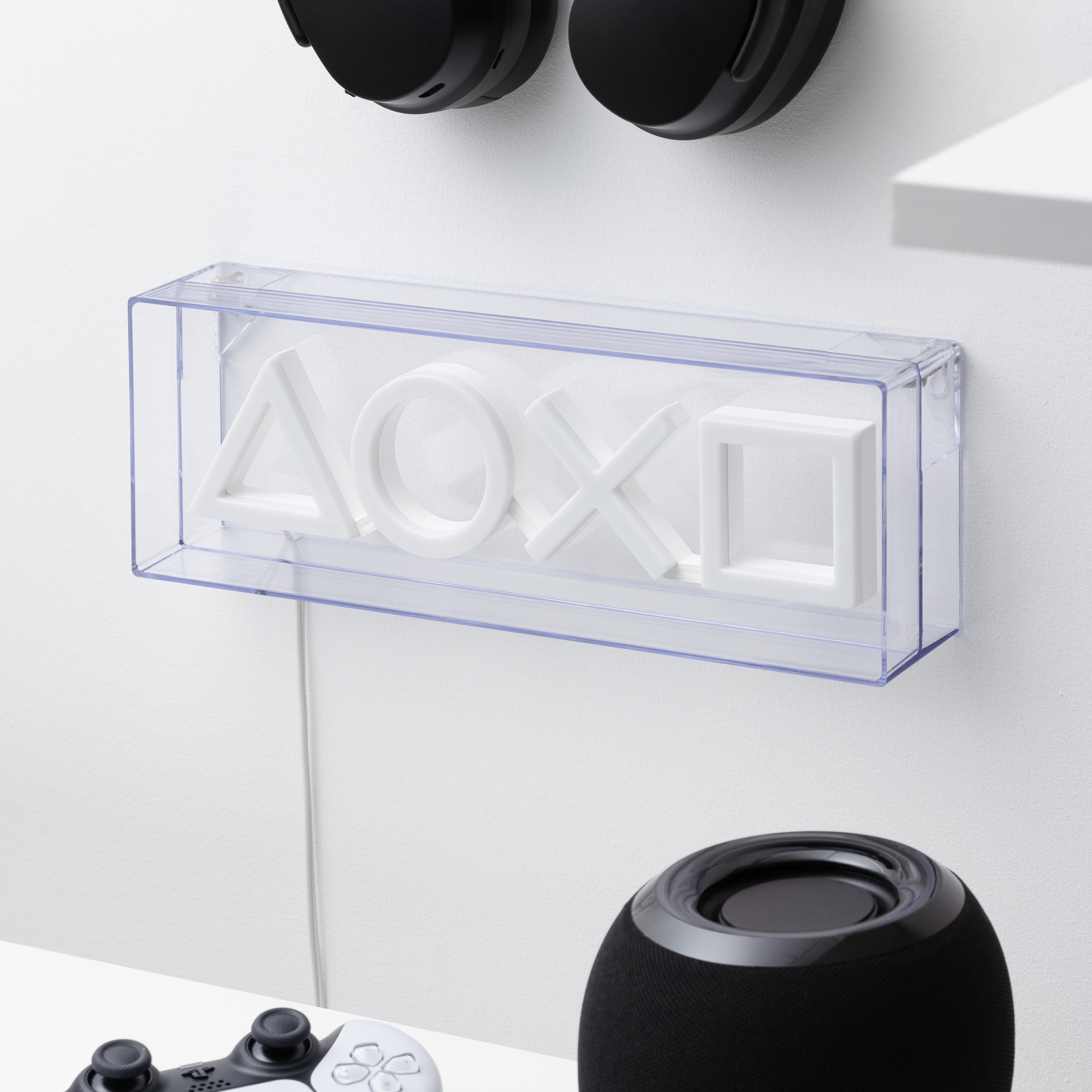 Eine transparente Plastikbox mit weißen PlayStation-Symbolen an einer weißen Wand. Darunter ein Lautsprecher und ein Controller.