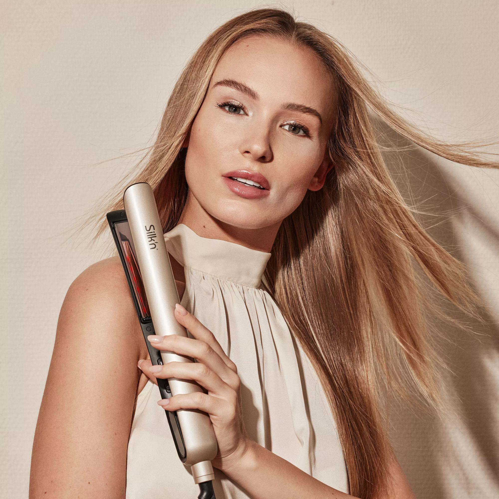 Kobieta trzymająca złotą prostownicę do włosów. Ma długie blond włosy i kremowy top.