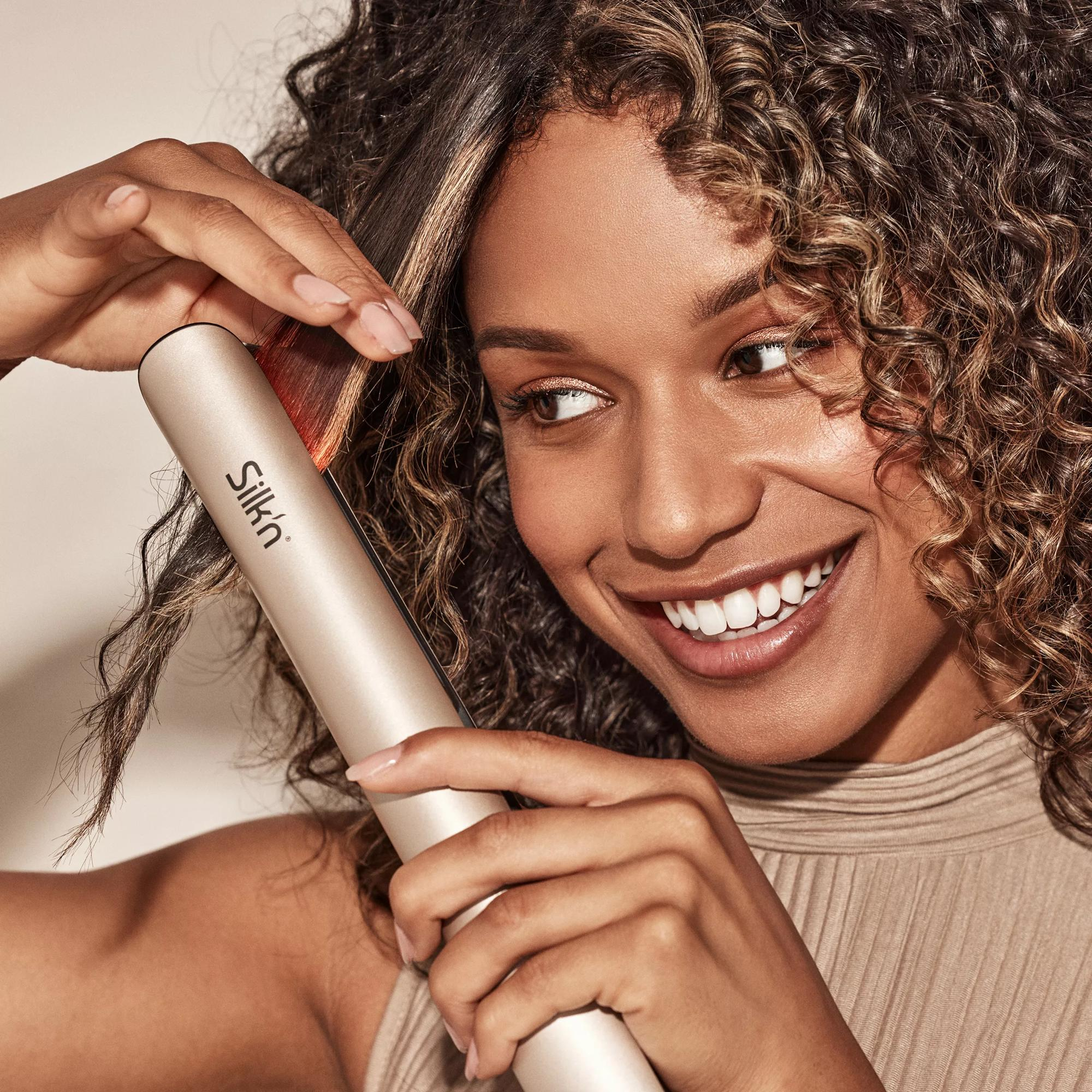 Kobieta uśmiecha się, używając prostownicy. Jej włosy są kręcone, a prostownica złota.