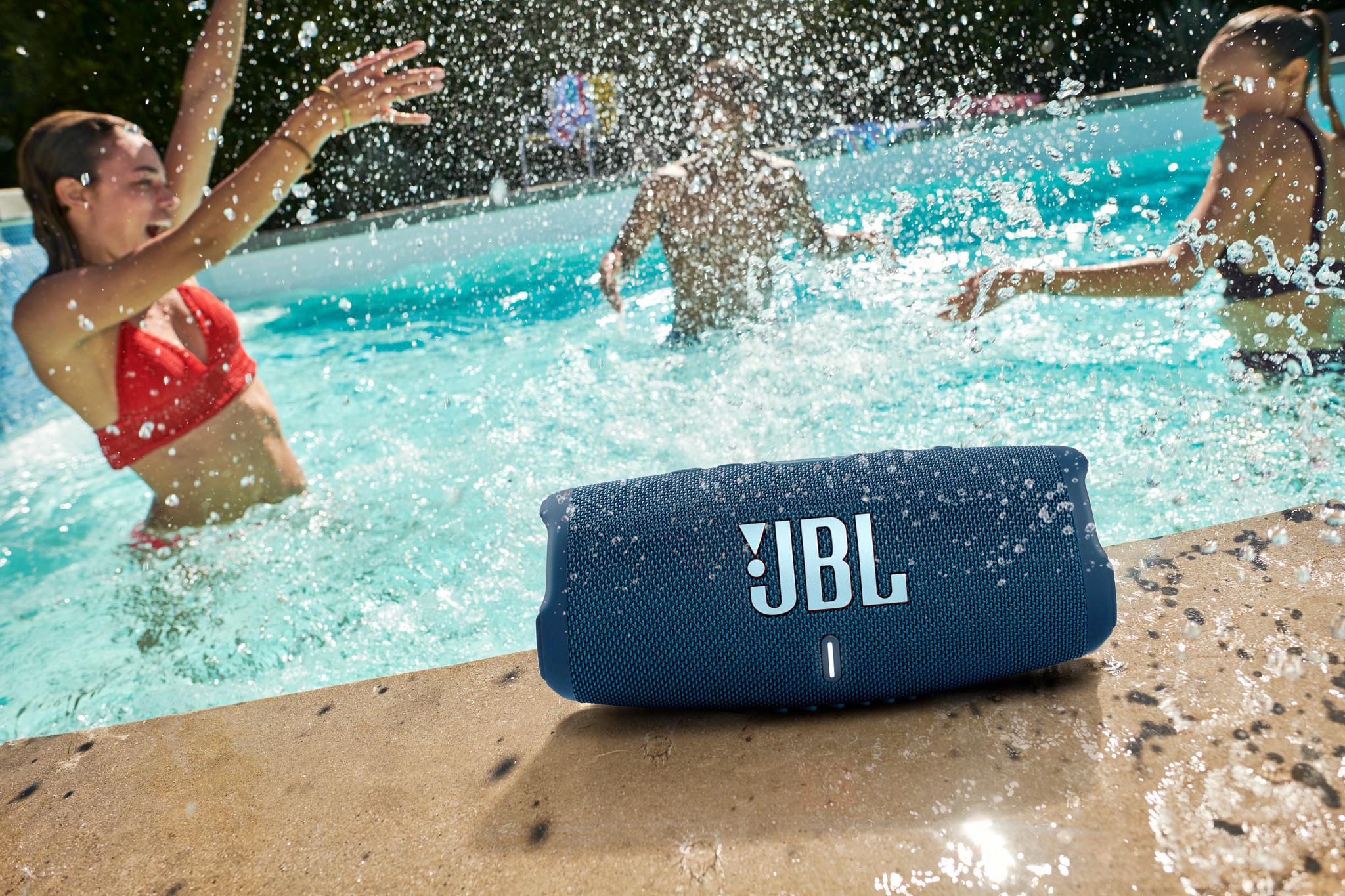 Ein blauer JBL-Lautsprecher am Poolrand mit spielenden Personen im Wasser.