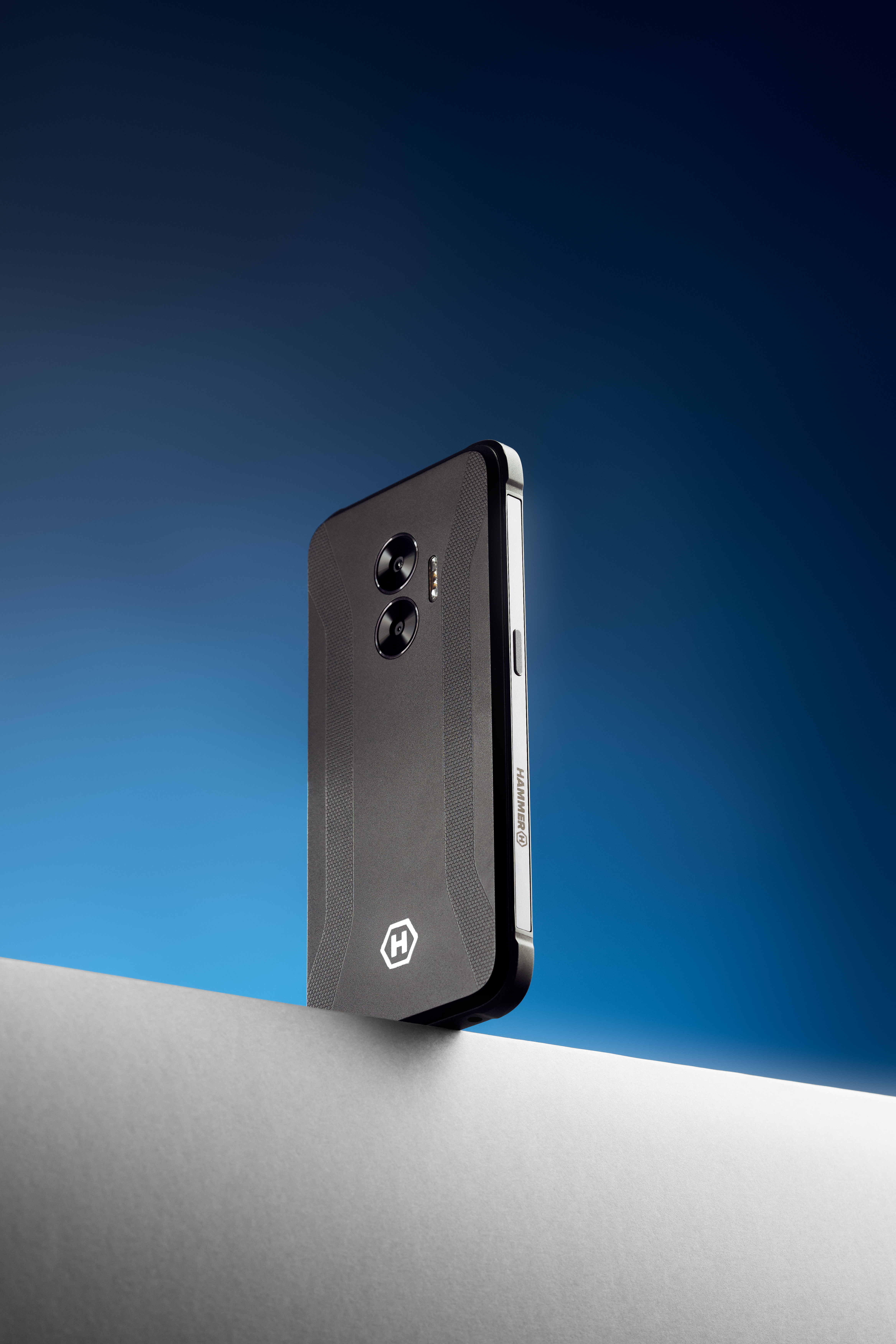 Czarny telefon stojący na białej powierzchni, z niebieskim tłem. Telefon jest ustawiony pod kątem.