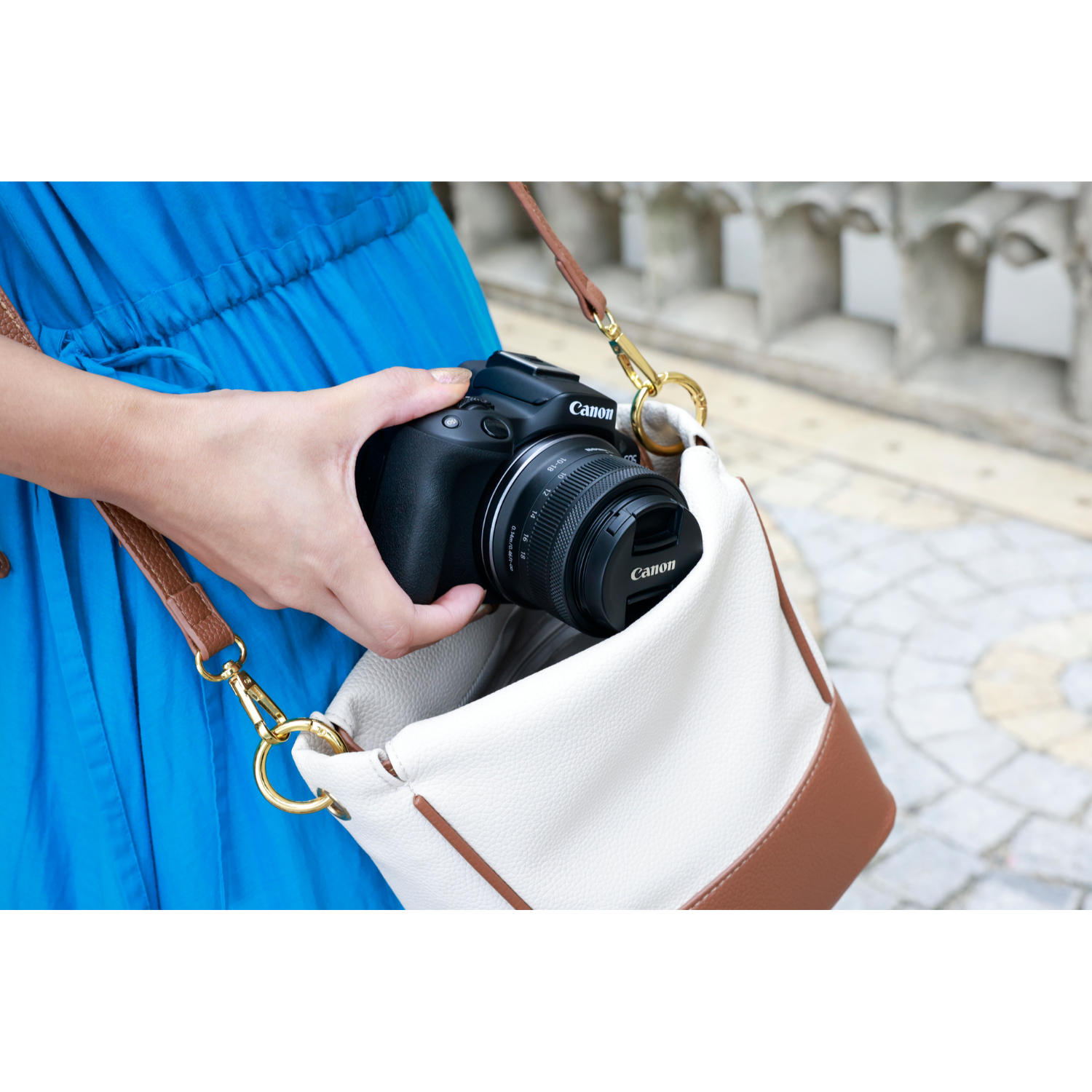 A woman holds a black Canon camera in a white and brown purse.