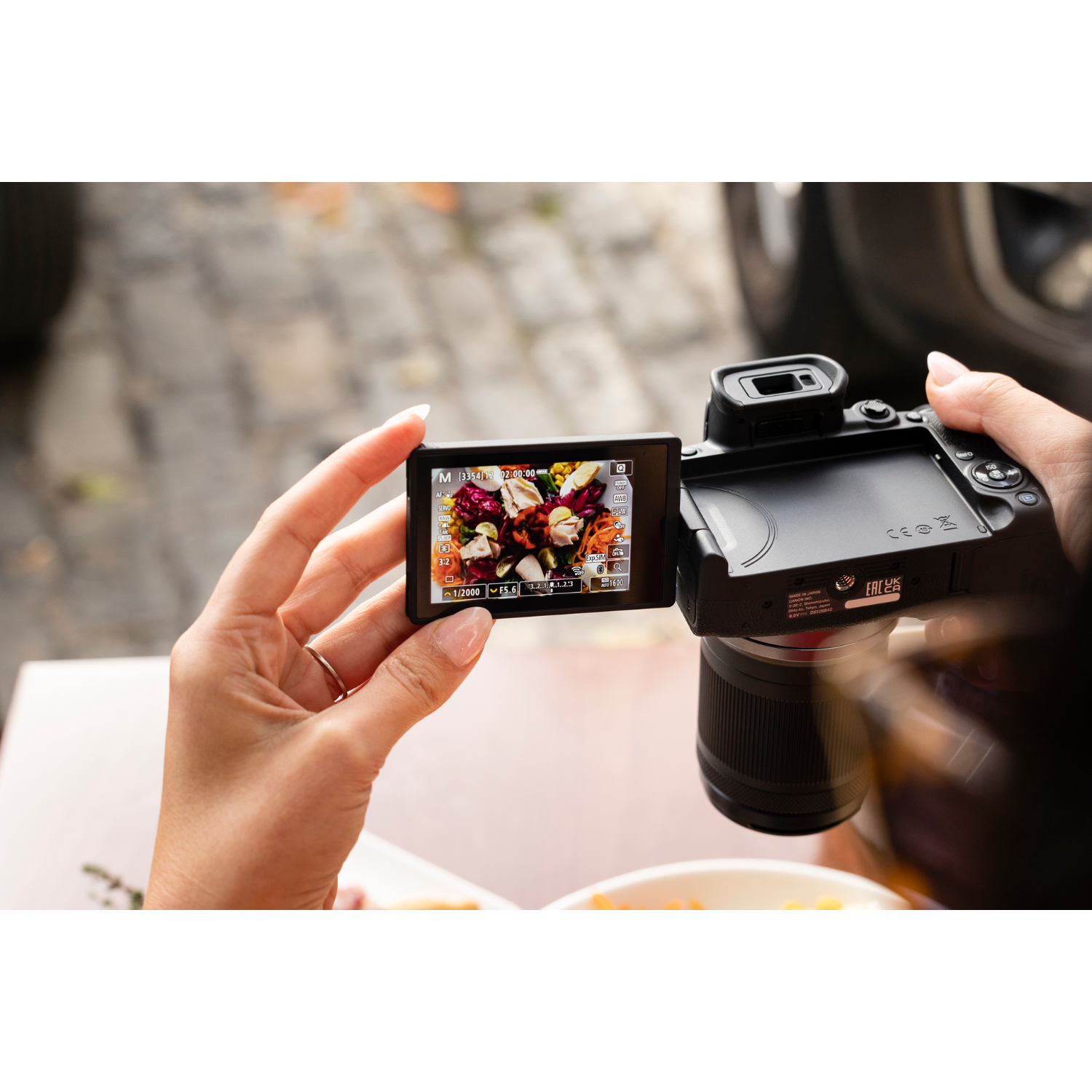 A woman holds a camera, viewing a colorful floral arrangement on the screen. The scene is set outdoors, with a table.