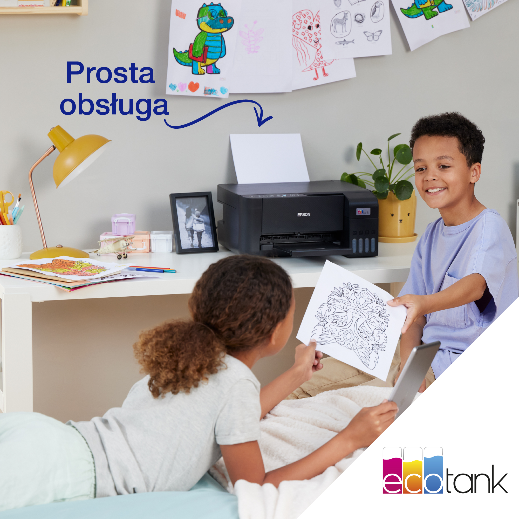 Dwoje dzieci przy biurku, jedno leży, drugie się uśmiecha; drukarka, rysunki, tablet.