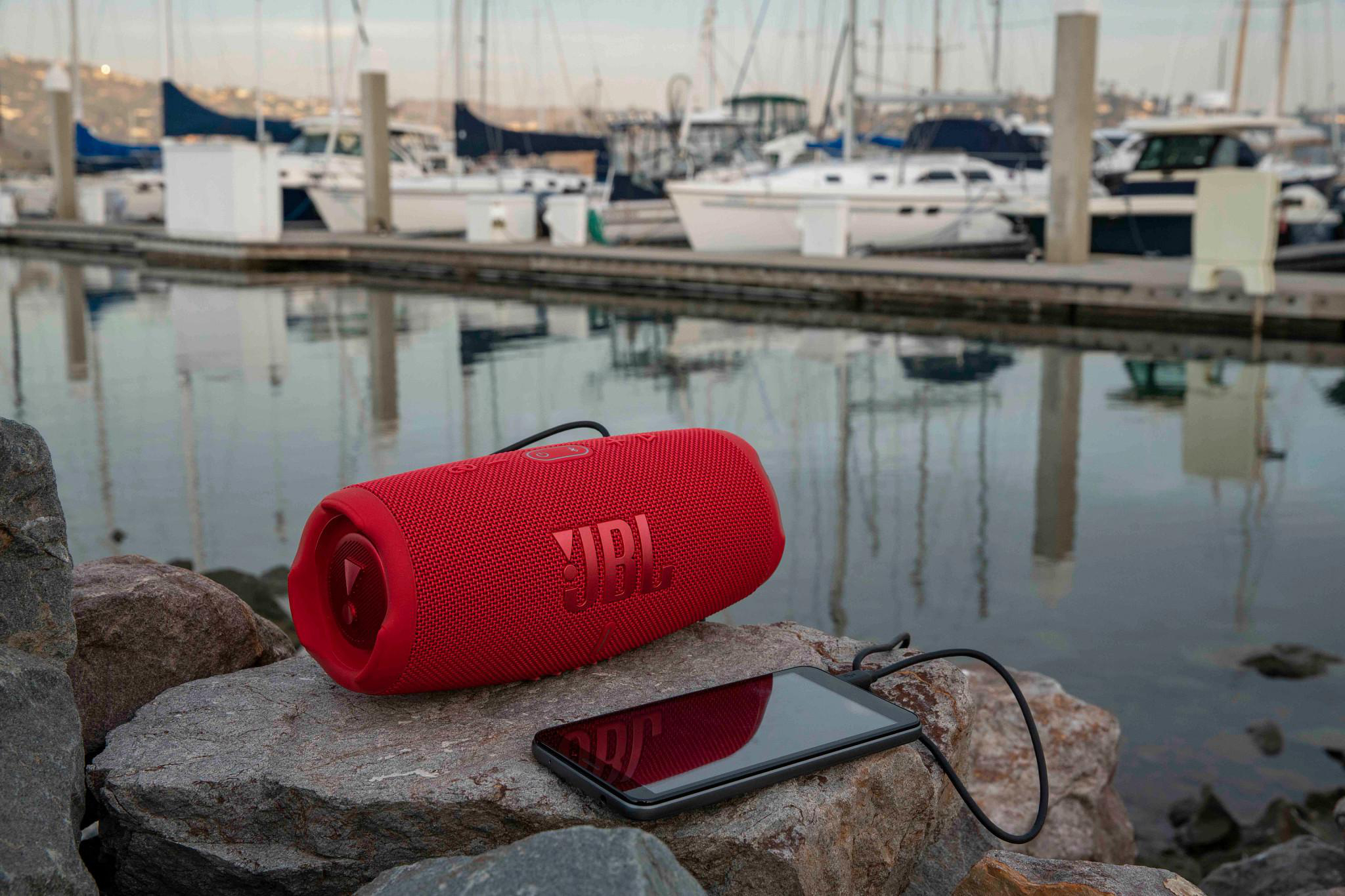 Ein roter JBL-Lautsprecher und ein Smartphone liegen auf Felsen am Wasser, Boote im Hintergrund.