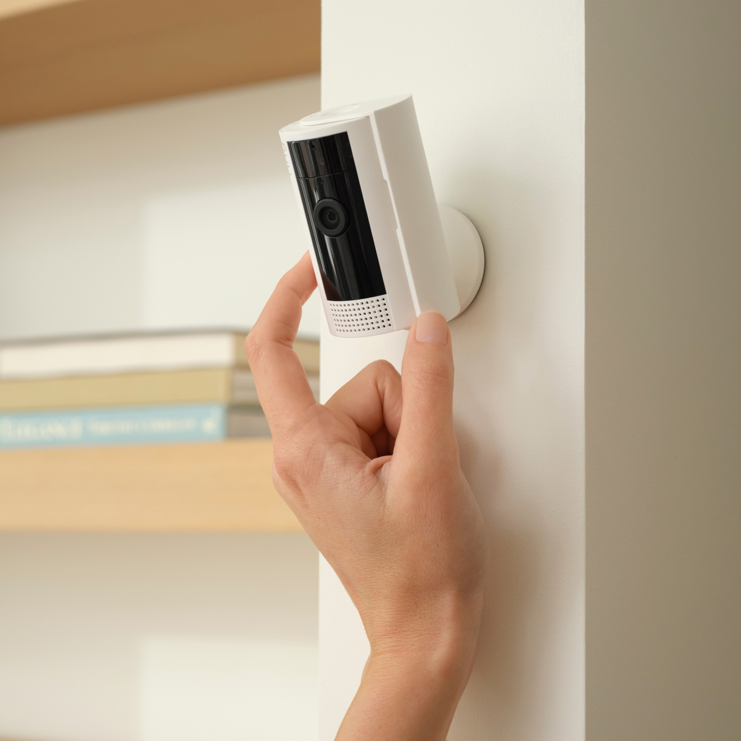 A person installs a white security camera on a white wall, near a wooden shelf.