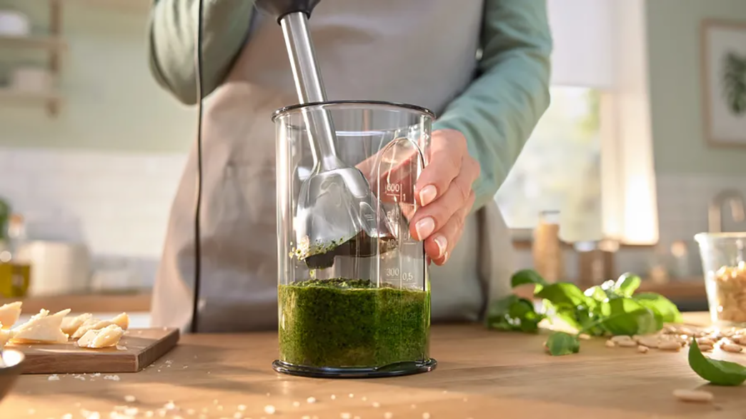 Egy személy kézi mixerrel zöld pestót kever üvegben.
