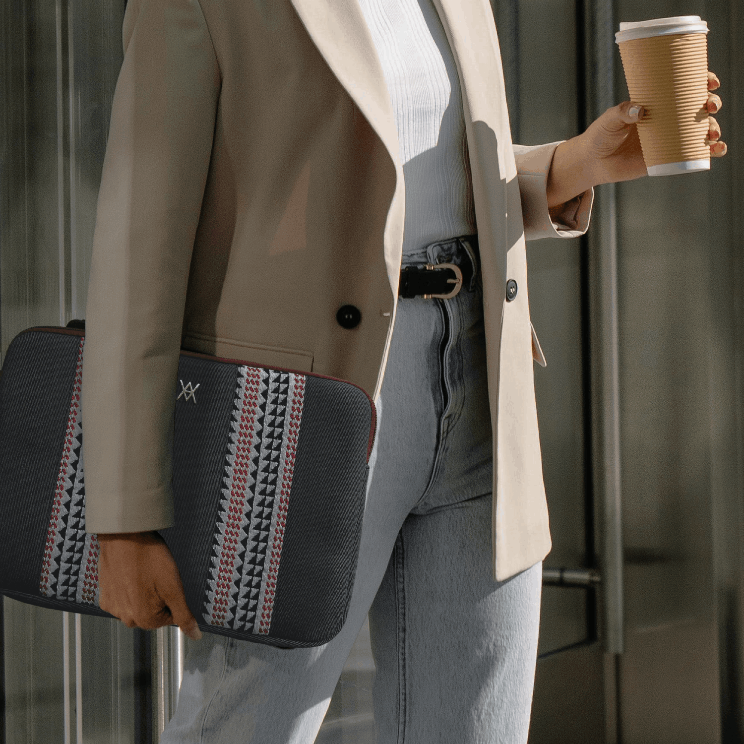 Mujer con blazer sostiene funda de portátil y café.