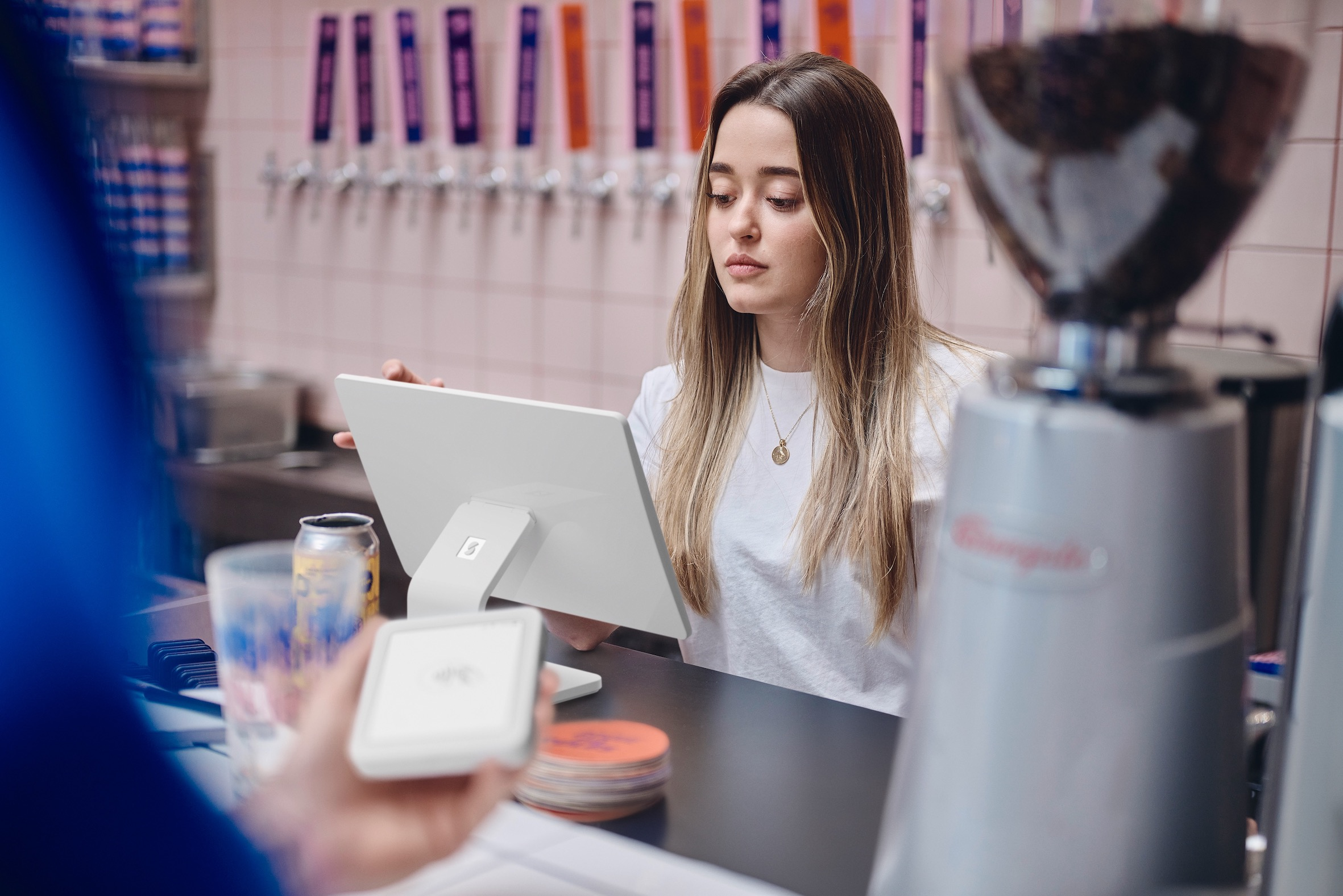 Una mujer en un mostrador usa una tableta. Una cafetera y grifos están en el fondo. Una persona con un lector de tarjetas.