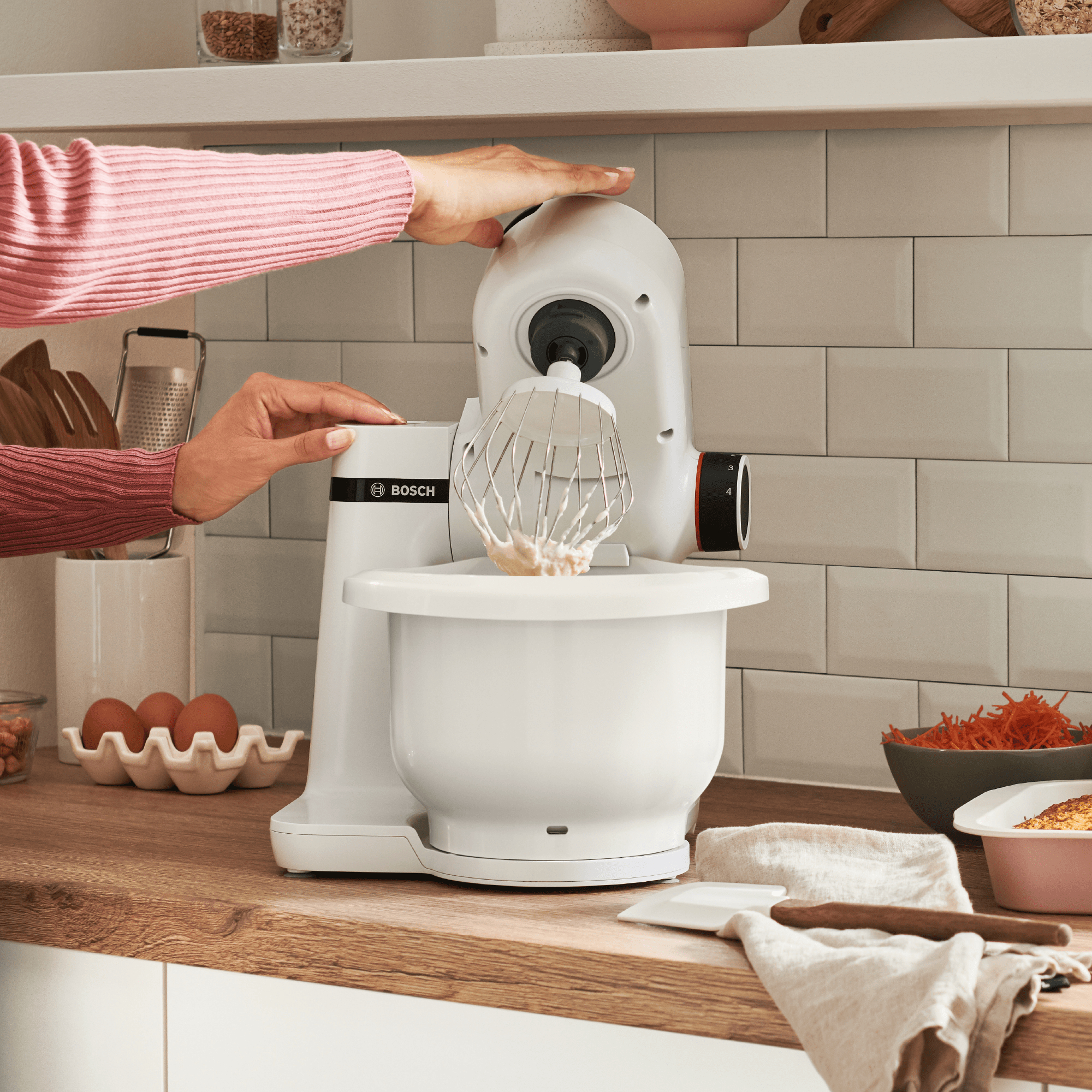 Frau benutzt einen weißen Küchenmixer mit Schneebesenaufsatz, mit Küchenartikeln und Essen auf der Theke.