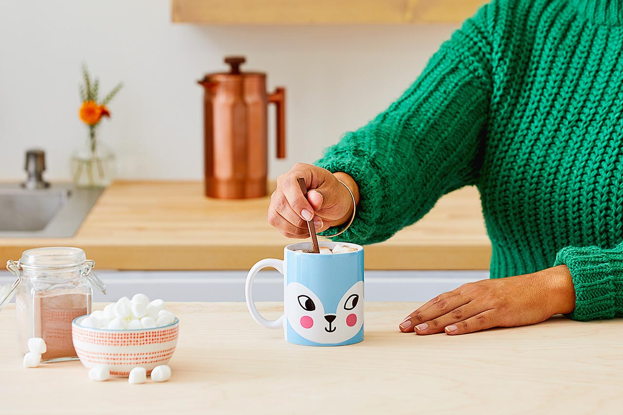 Eine Person rührt Kakao in einer blau-fuchsgesichtigen Tasse um. Marshmallows und Kakaopulver stehen daneben. Ein Kupferkessel ist im Hintergrund.