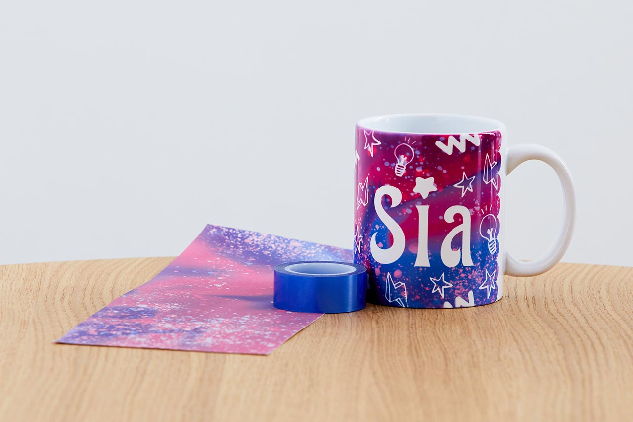 Eine Tasse mit "Sia", neben einem Blatt Papier und einer Kleberolle, alles auf einem Holztisch.