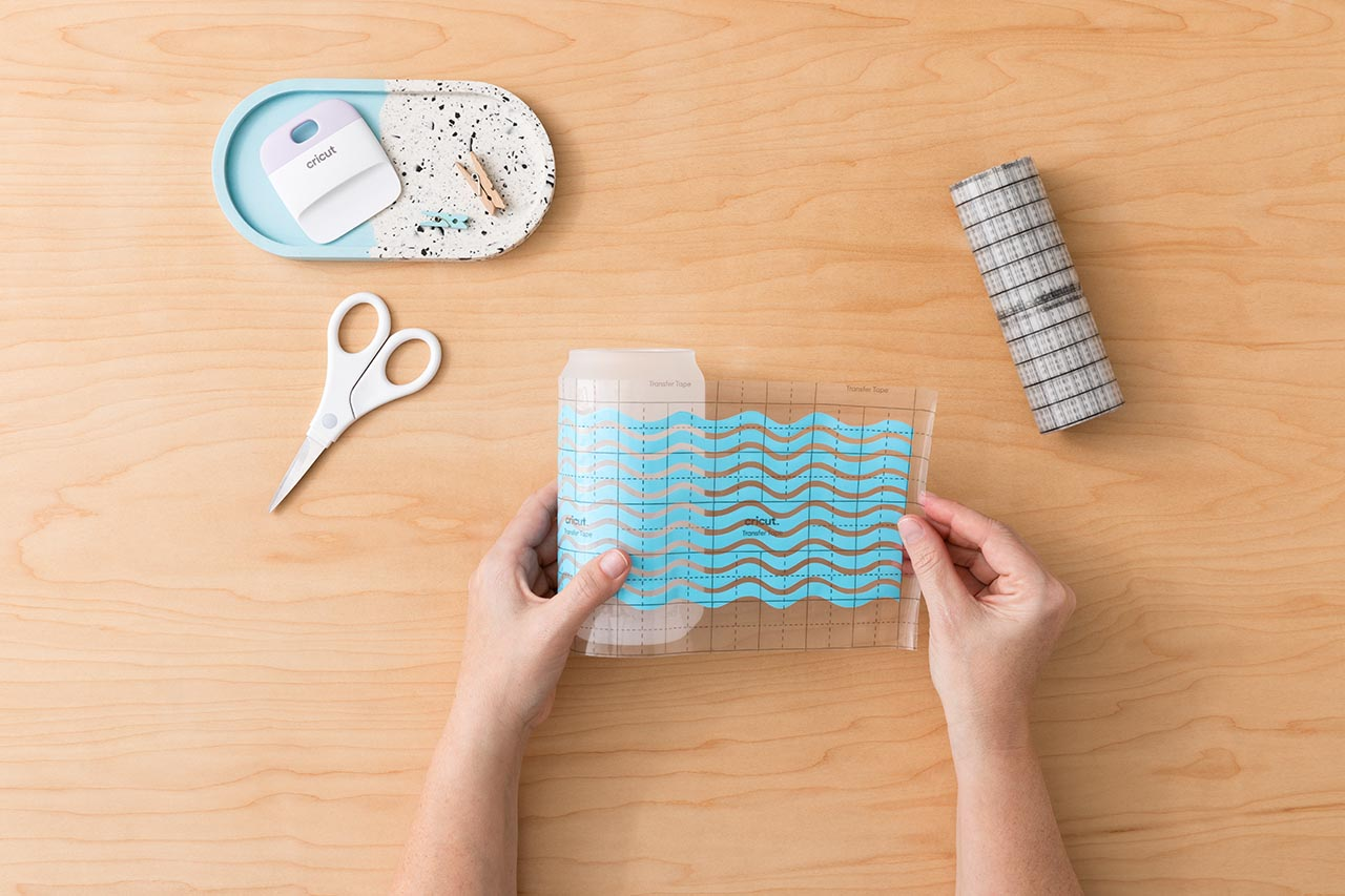 Hände halten Cricut-Transferband mit Wellendesign, Schere, eine Ablage mit einem Schaber und eine Rolle gemustertes Klebeband auf Holz.