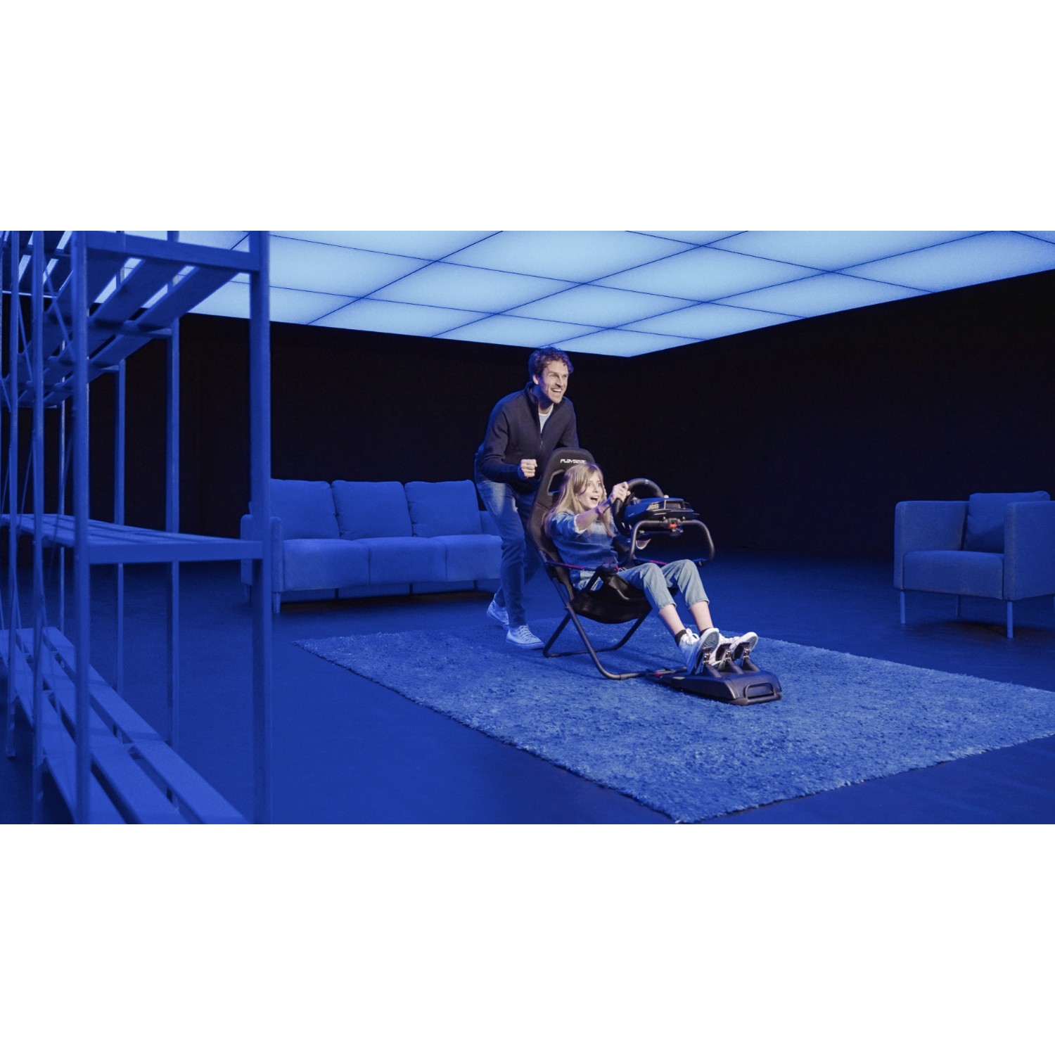 A man pushes a girl in a racing simulator chair in a blue-lit room with shelves.