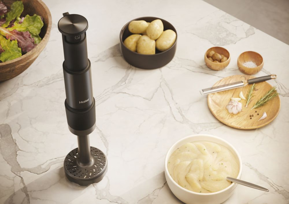 Siyah bir Haier el blenderi ve patates, sarımsak ve salata gibi malzemeler mermer bir yüzey üzerinde.