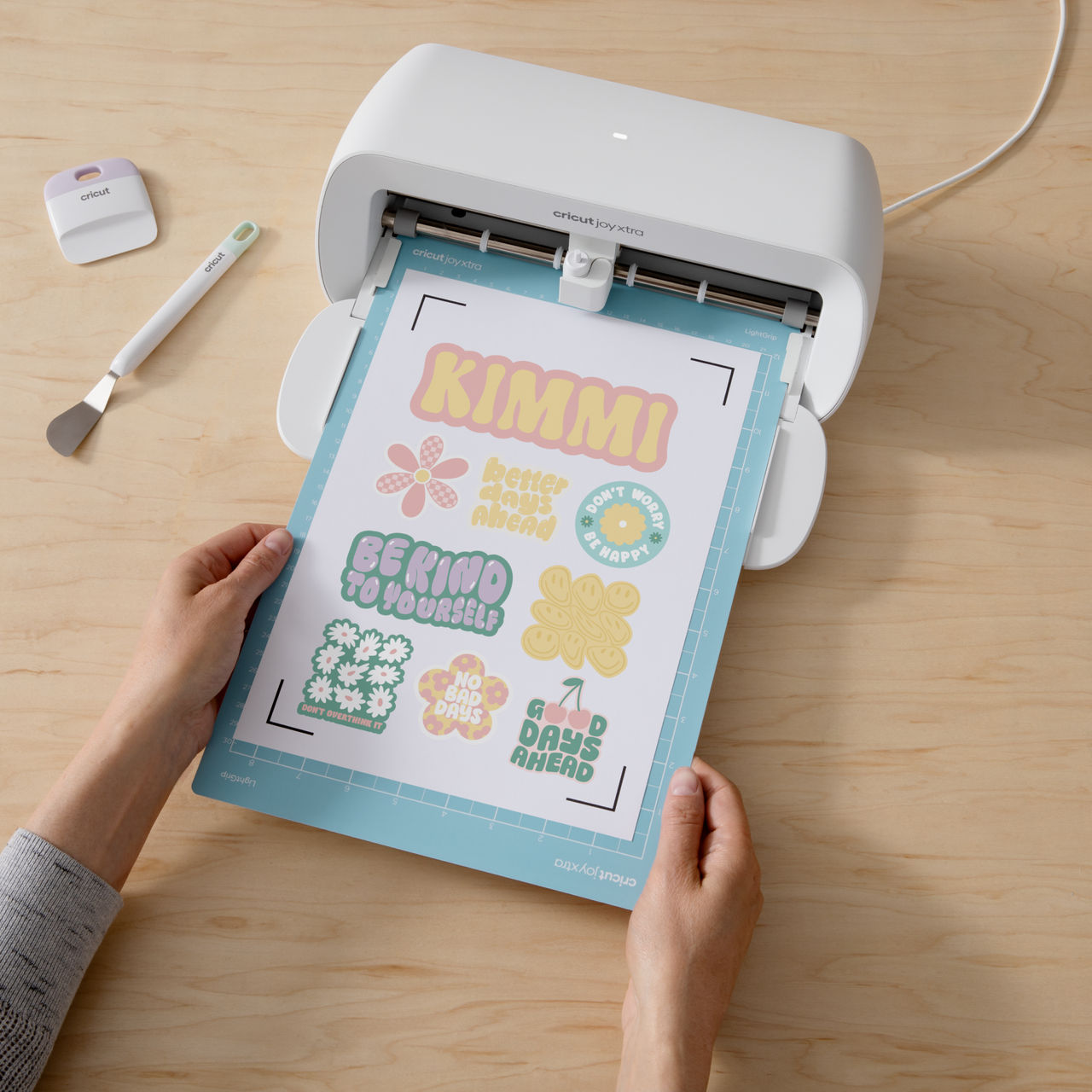 Hände halten eine Cricut Joy Xtra Maschine mit einem geschnittenen Bogen bunter Aufkleber.