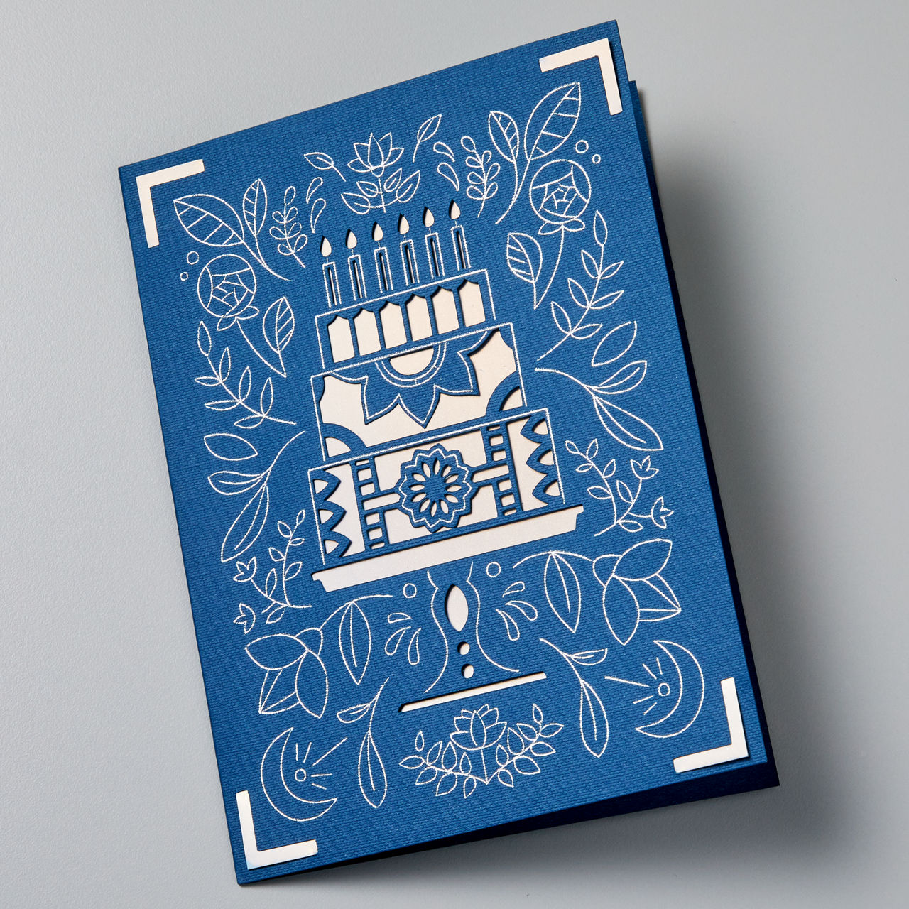 Eine blaue Geburtstagskarte mit einem weißen Kuchen-Design, umgeben von weißen Blumen- und Blättermustern.
