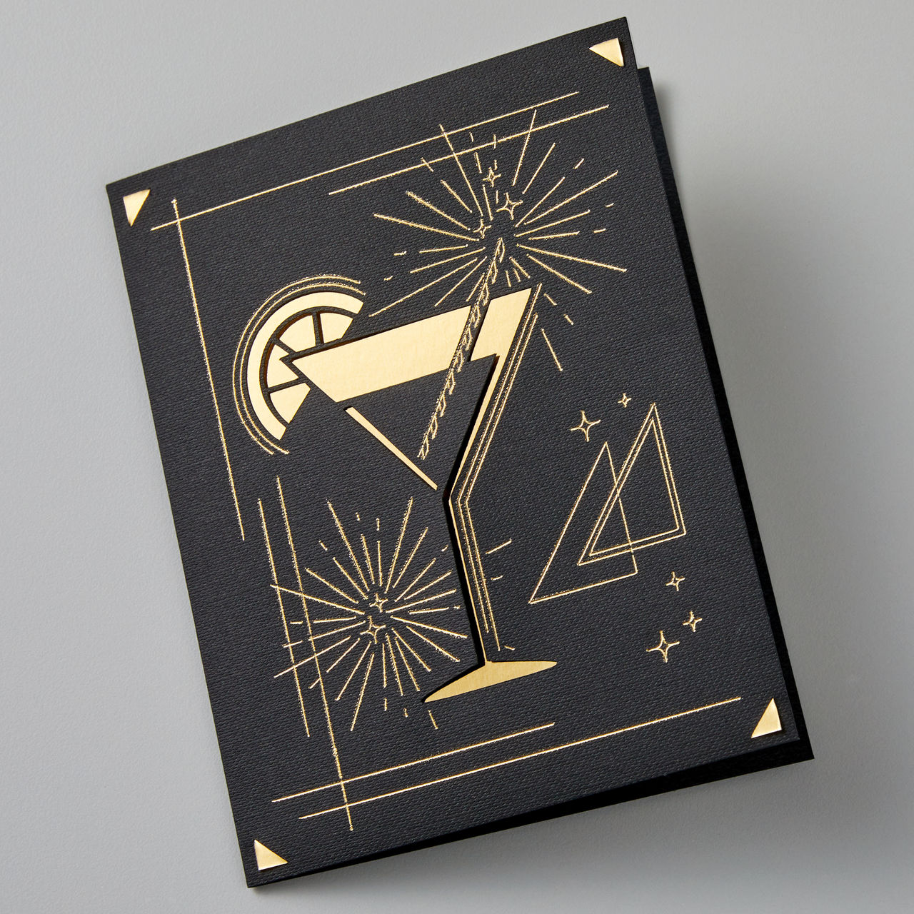 Eine schwarze Karte mit einem goldenen Cocktail-Design. Sie hat geometrische Formen und Feuerwerksmuster.