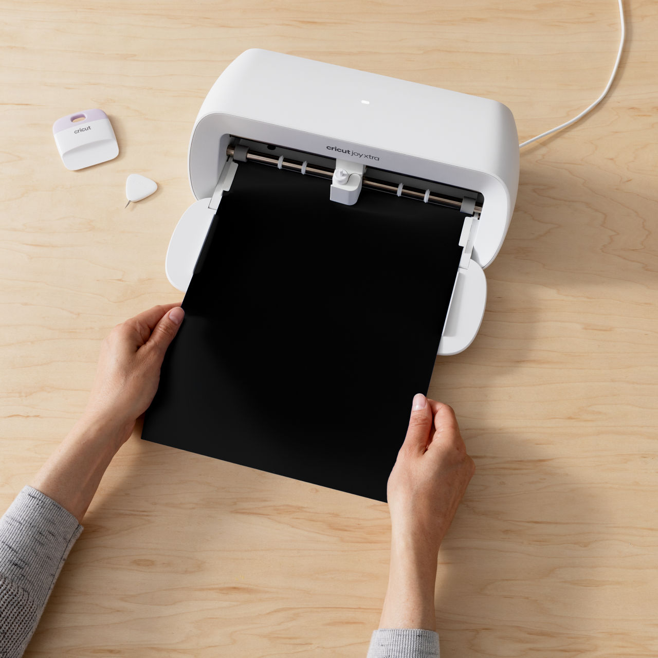Hände halten schwarzes Papier in einer weißen Cricut Joy Xtra Maschine. Holztisch.