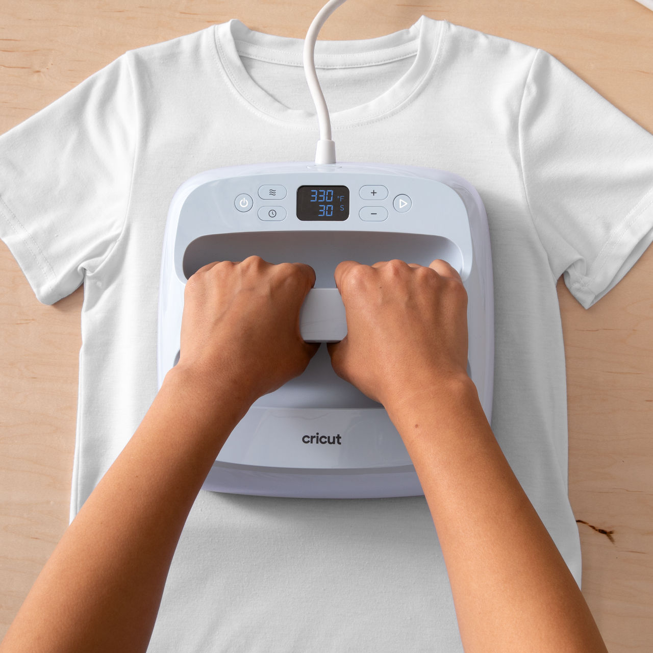 Hände, die eine weiße Cricut-Heizpresse auf einem weißen T-Shirt auf Holz verwenden.
