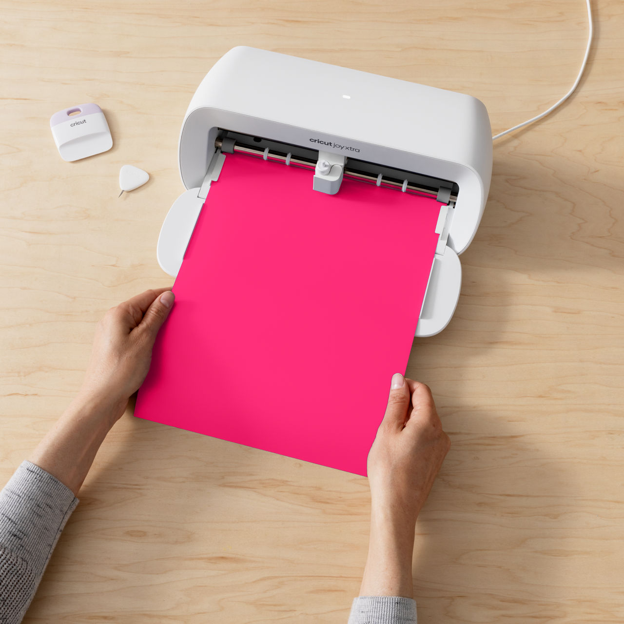 Eine weiße Cricut-Maschine mit einem rosa Blatt Papier, das hineingeführt wird. Hände halten das Papier.