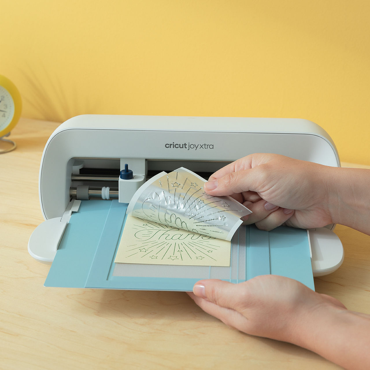 Hände ziehen ein Design von einer Schneidematte ab. Eine Cricut Joy Xtra Maschine und gelber Hintergrund sind sichtbar.