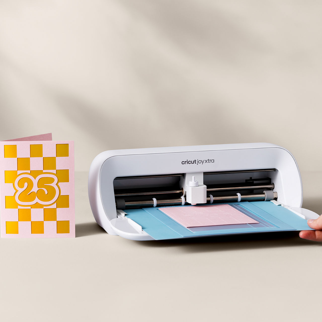 Eine Cricut Joy Xtra Maschine schneidet eine Karte. Eine fertige Karte liegt daneben. Der Hintergrund ist neutral.