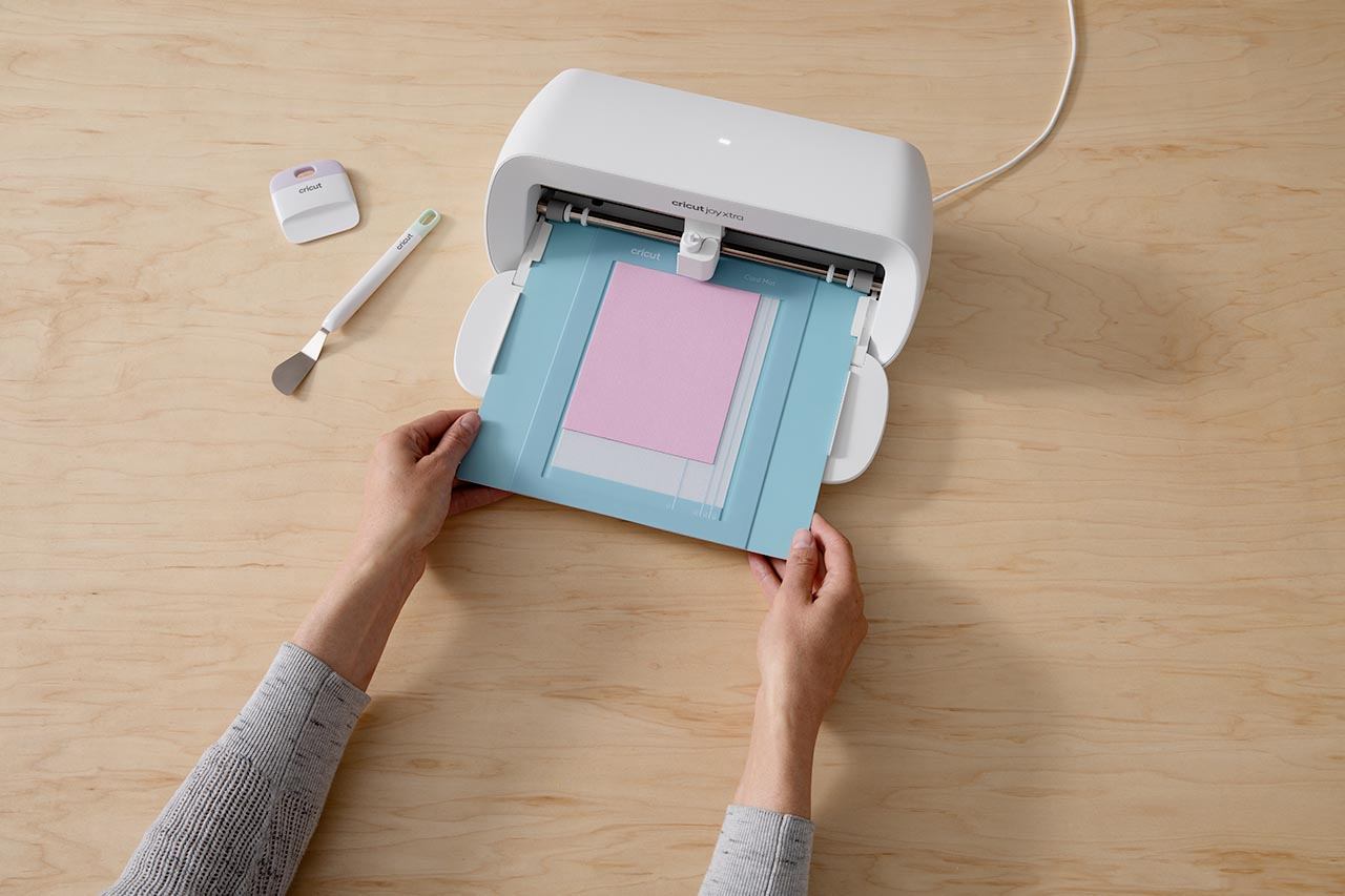 Hände, die eine Schneidematte in eine Cricut Joy Xtra Maschine legen. Holzoberfläche und Werkzeuge sind sichtbar.