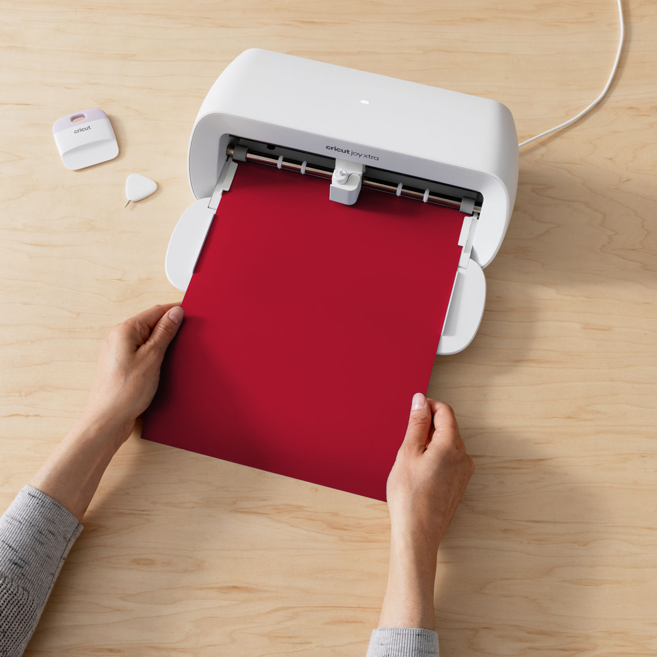 Eine weiße Cricut-Maschine mit einem roten Papier, das eingelegt wird. Hände halten das Papier auf einer Holzoberfläche.