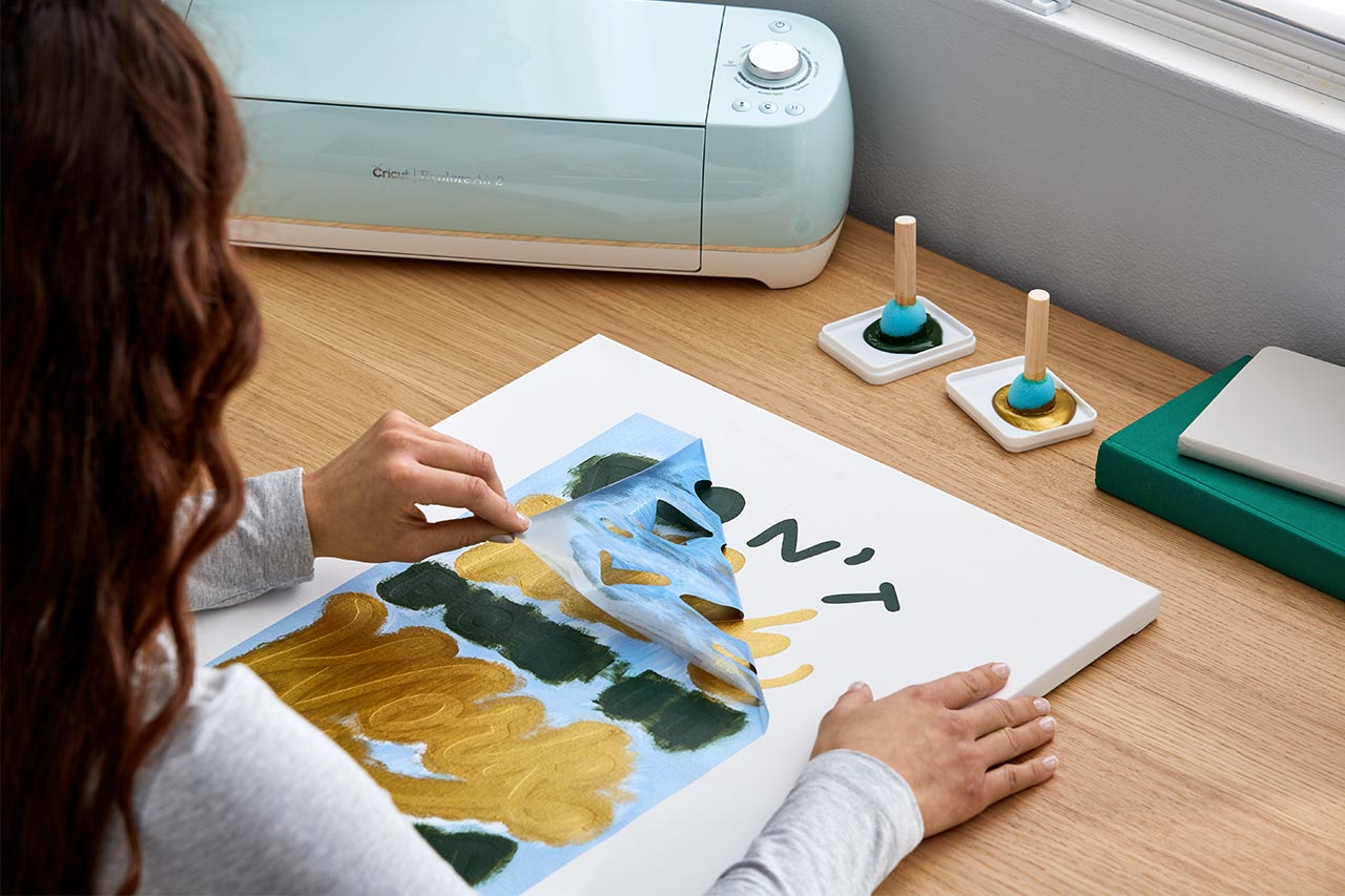 Person zieht Farbe von einer Leinwand ab, Cricut-Maschine im Hintergrund, kleine Dekoration.