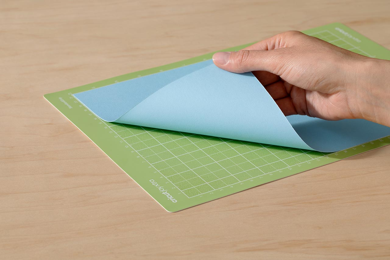 Eine Hand, die ein blaues Material von einer grünen Schneidematte mit 'Cricut Joy Xtra' abzieht.