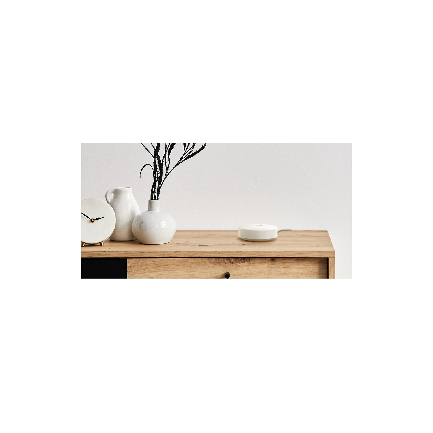 Un meuble en bois avec un vase blanc avec des branches de plante, une horloge blanche et un appareil domestique intelligent blanc dessus.