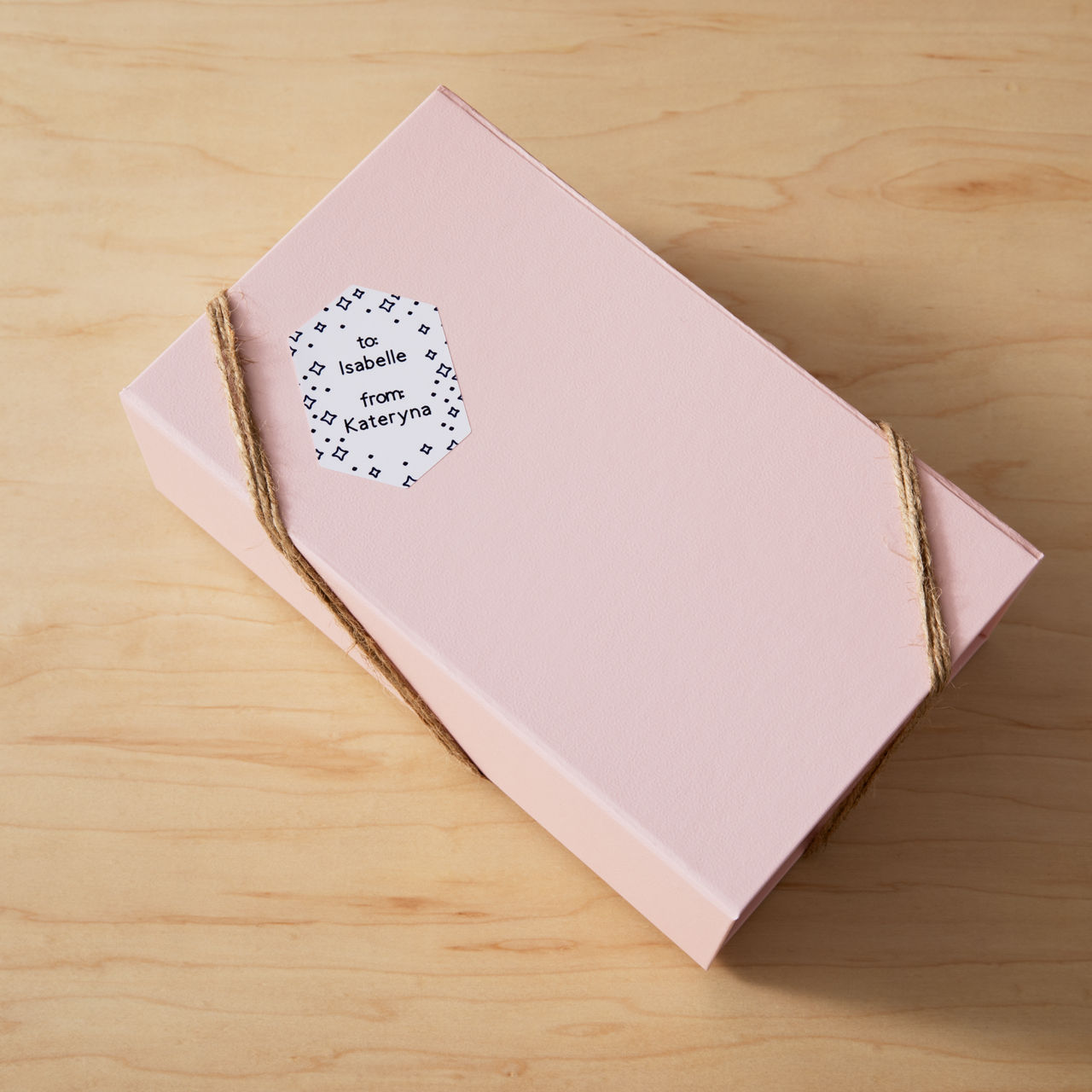 Rosa Geschenkbox mit Bindfaden, beschriftet 'To Isabelle from Kateryna' auf Holzoberfläche.