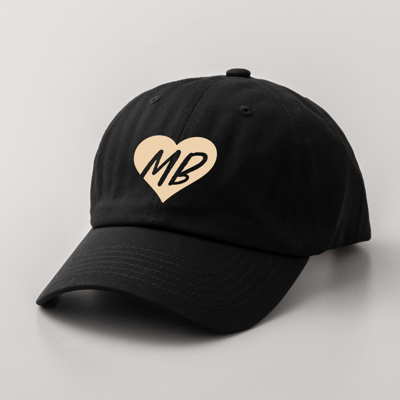 Schwarze Baseballkappe mit einem hellbraunen Herz mit den Buchstaben MB. Die Kappe liegt auf grauem Hintergrund.