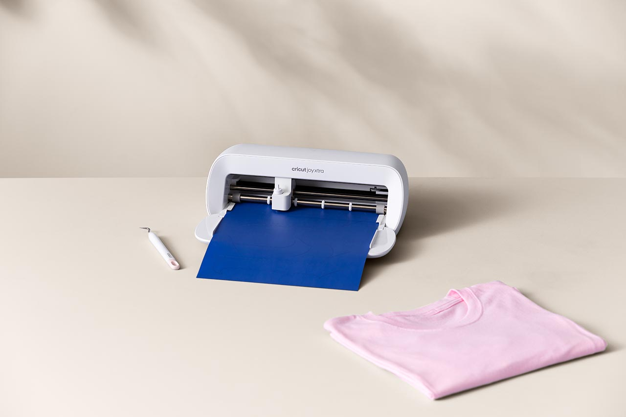 Eine weiße Cricut Joy Xtra Maschine mit blauem Papier und einem pinkfarbenen T-Shirt auf beigem Untergrund.